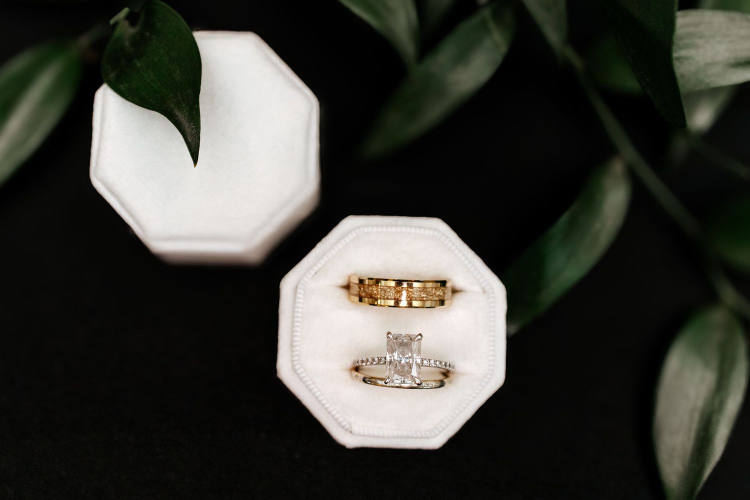 Gold wedding bands and diamond engagement ring displayed in a white velvet octagon ring box with green leaves.