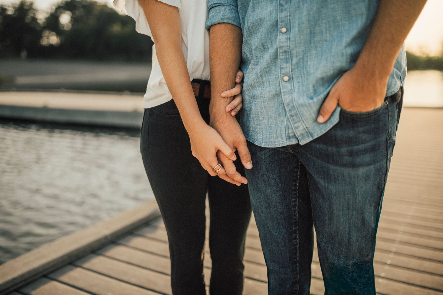 Close-up of couple holding hands on a lakeside dock at golden hour, wearing jeans and casual tops.