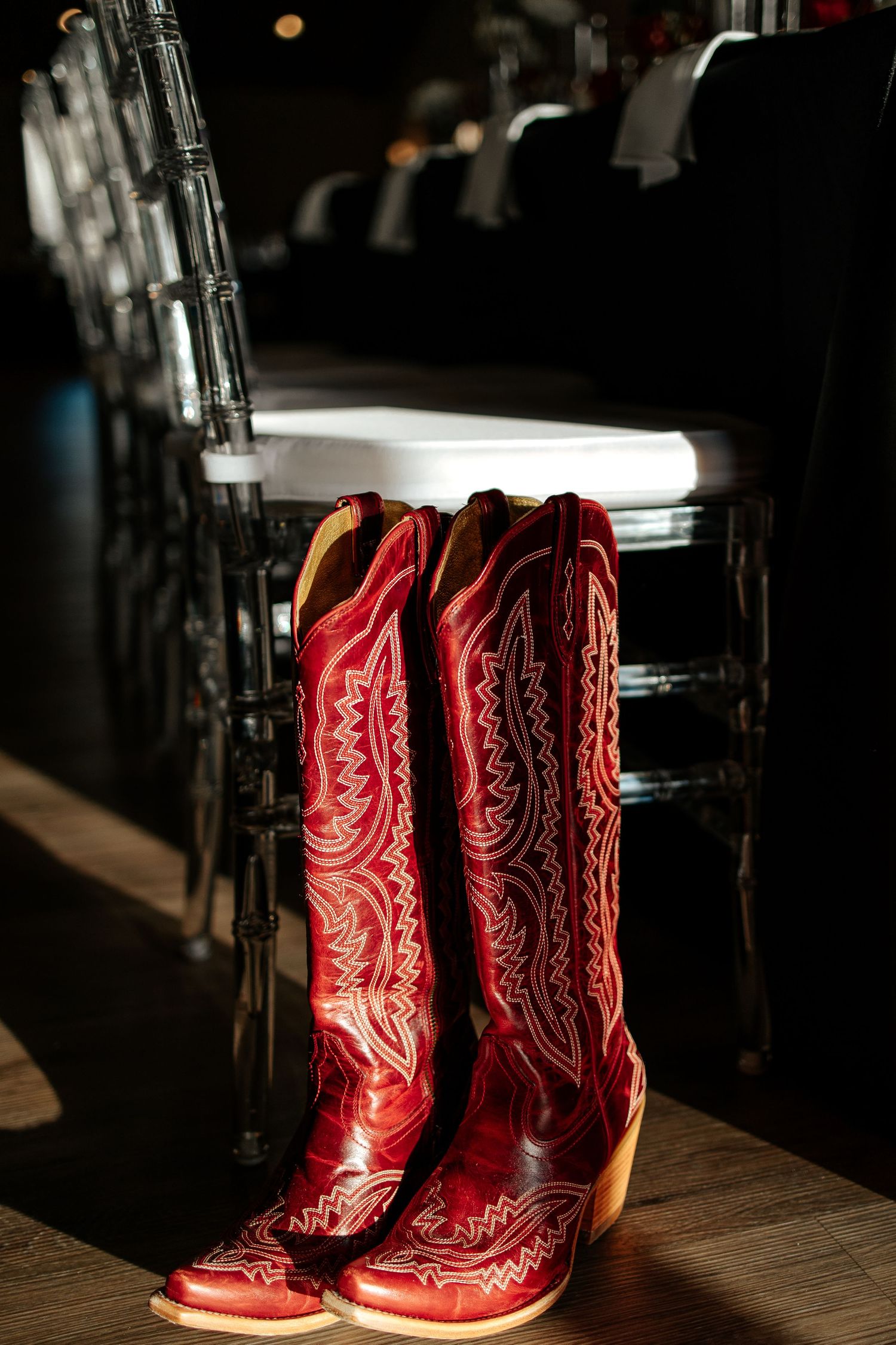 Red cowboy boots with white stitching stand on wooden floor beside black chairs at an elegant event venue.
