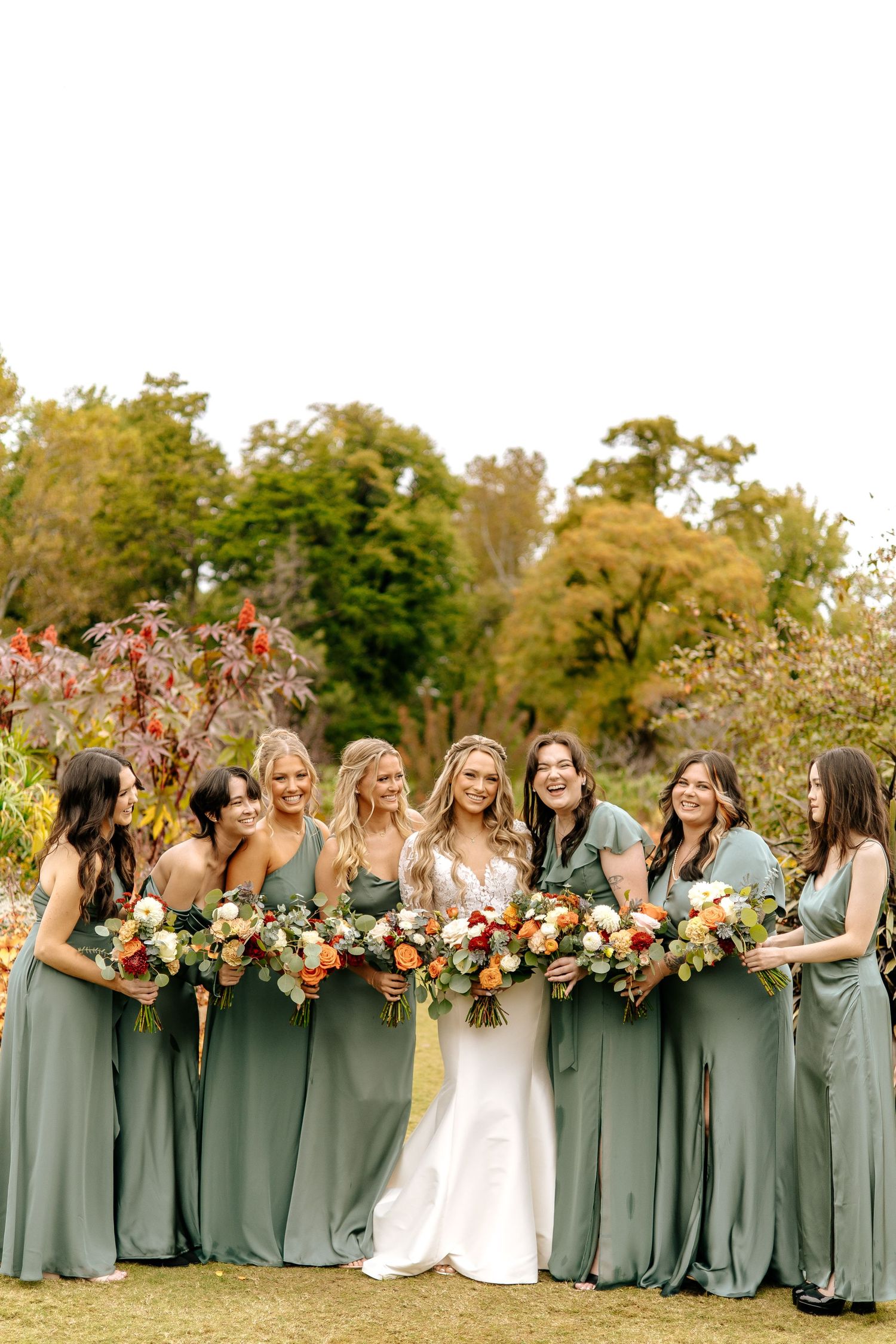 Bride in fitted white gown poses with seven bridesmaids in sage green dresses holding colorful autumn bouquets outdoors.