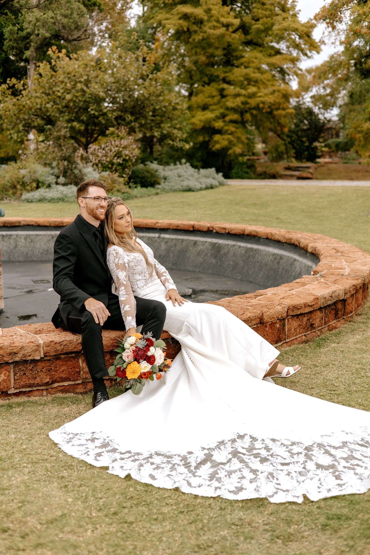 Bride and groom sit together on brick fountain edge in park, brides lace train spread across lawn.
