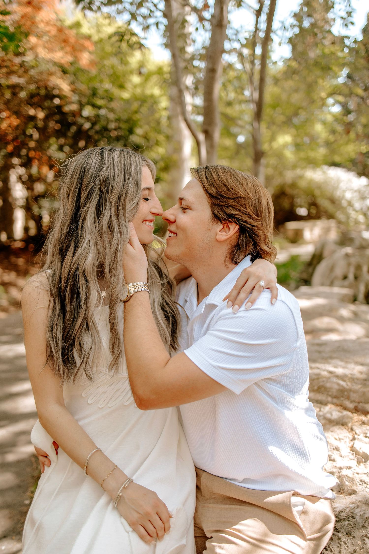 Happy couple sharing a romantic nose-to-nose moment outdoors, surrounded by lush green trees in soft sunlight.