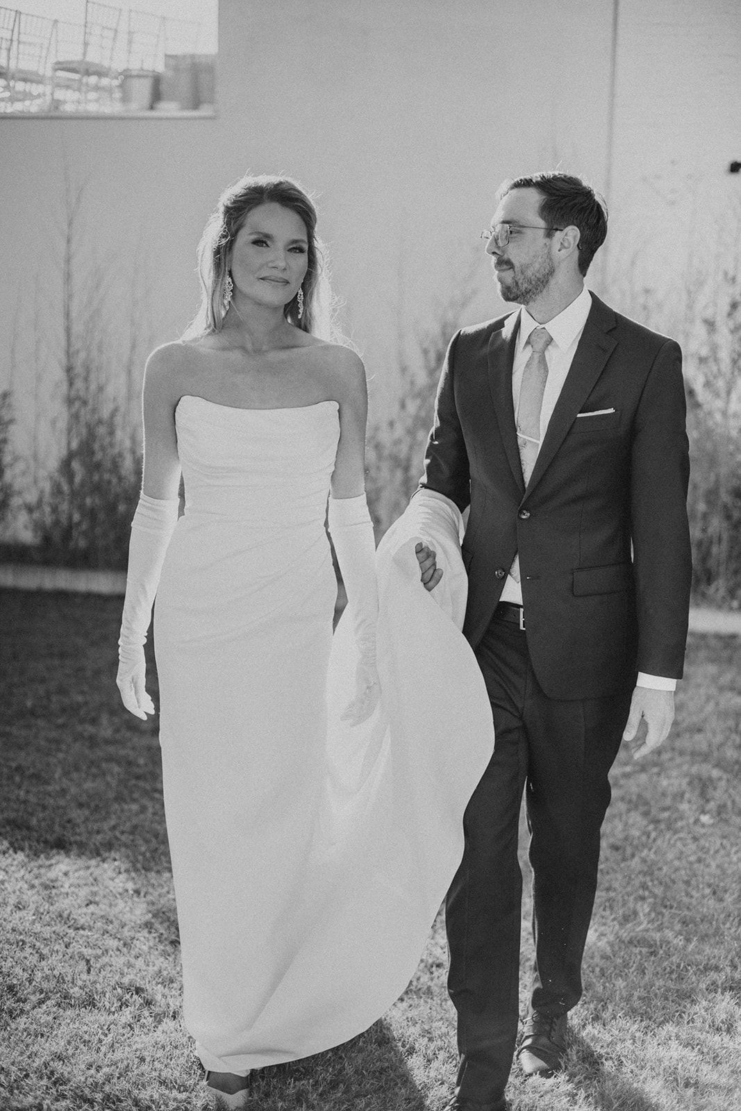 Black and white photo of a bride in a strapless gown with gloves and groom in a dark suit walking together.