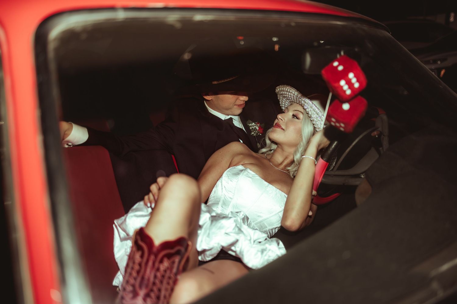 Bride and groom share a romantic moment inside a vintage red car at night.