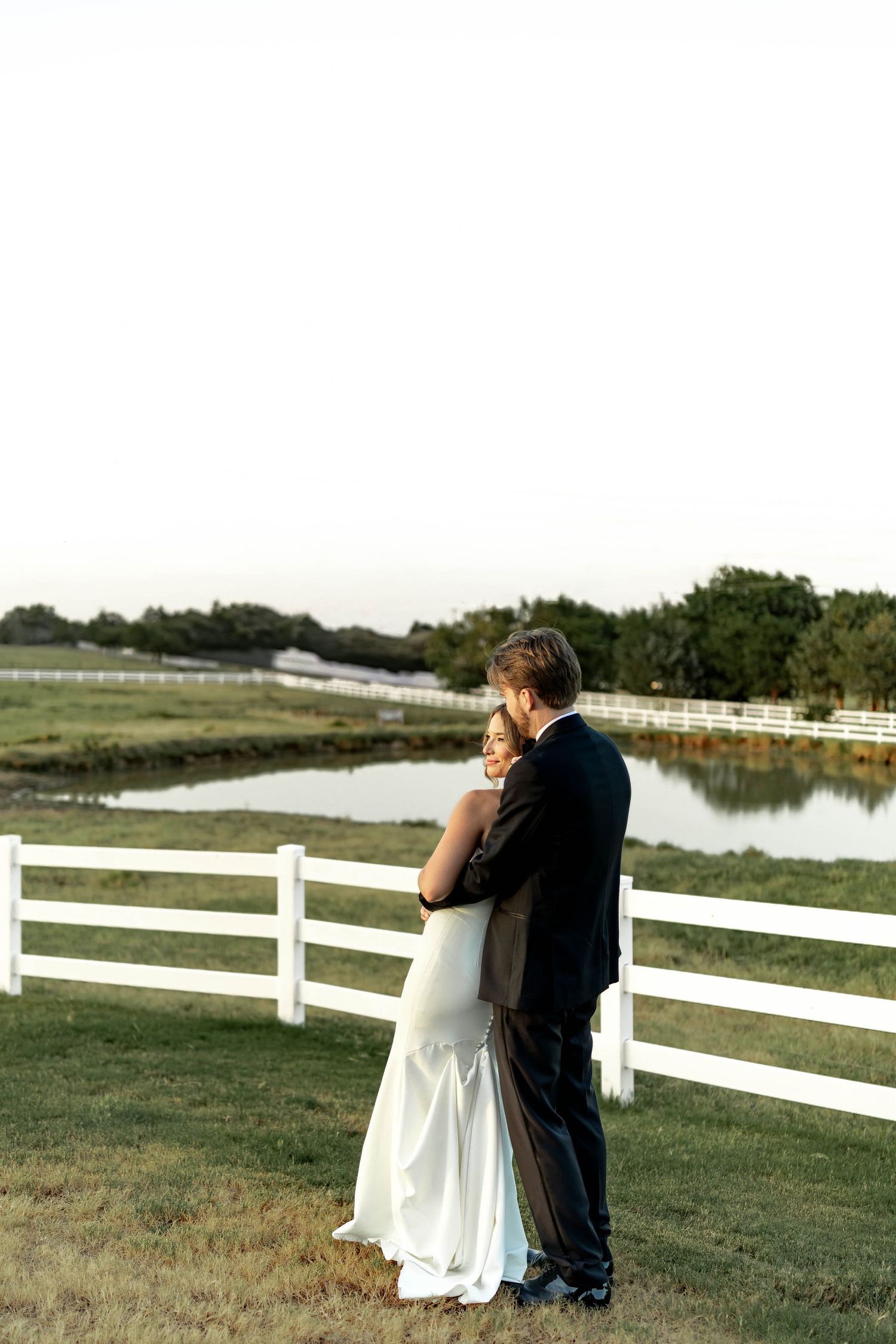 Bride and groom embracing near white fence by pond at golden hour outdoor wedding venue.