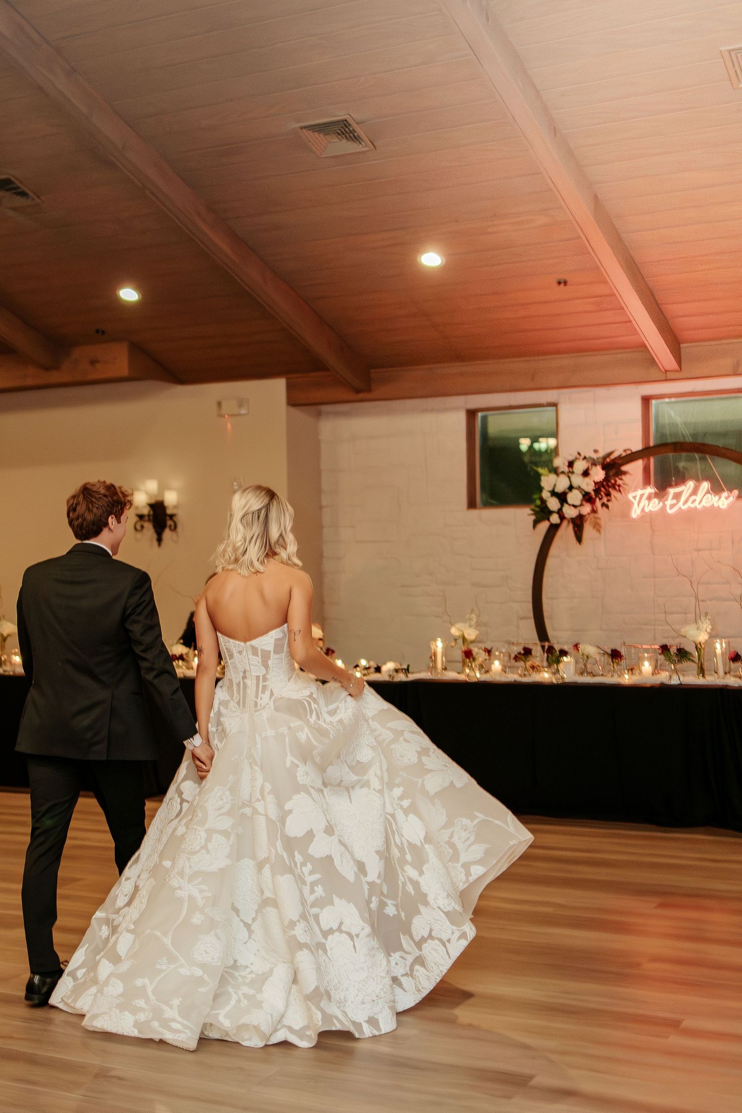 Bride and groom share first dance in elegant reception hall with floral arch and neon sign backdrop.
