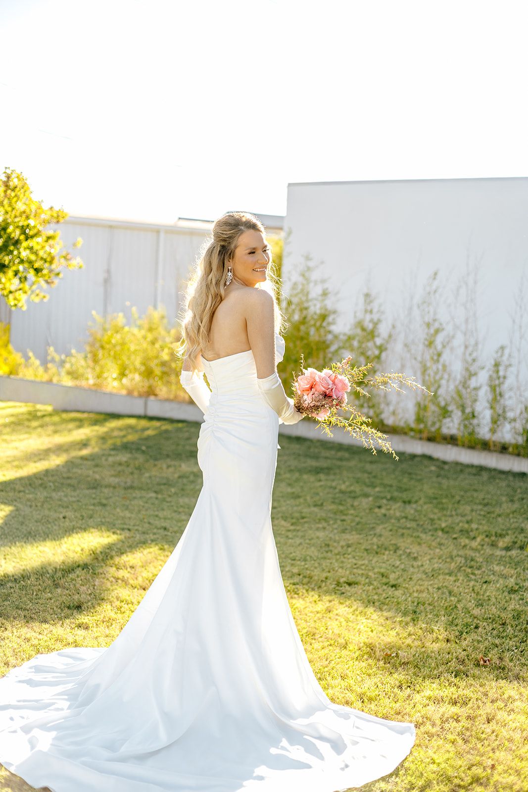 Bride in white strapless mermaid gown with train holds pink bouquet, posing outdoors on green lawn.