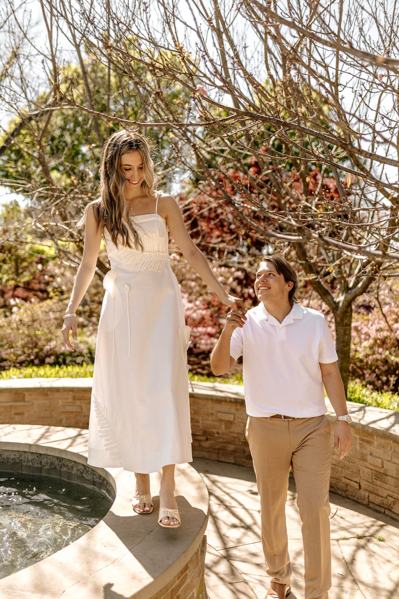 Couple holding hands by a garden fountain, woman in white dress standing on stone edge, man in white polo and khakis.