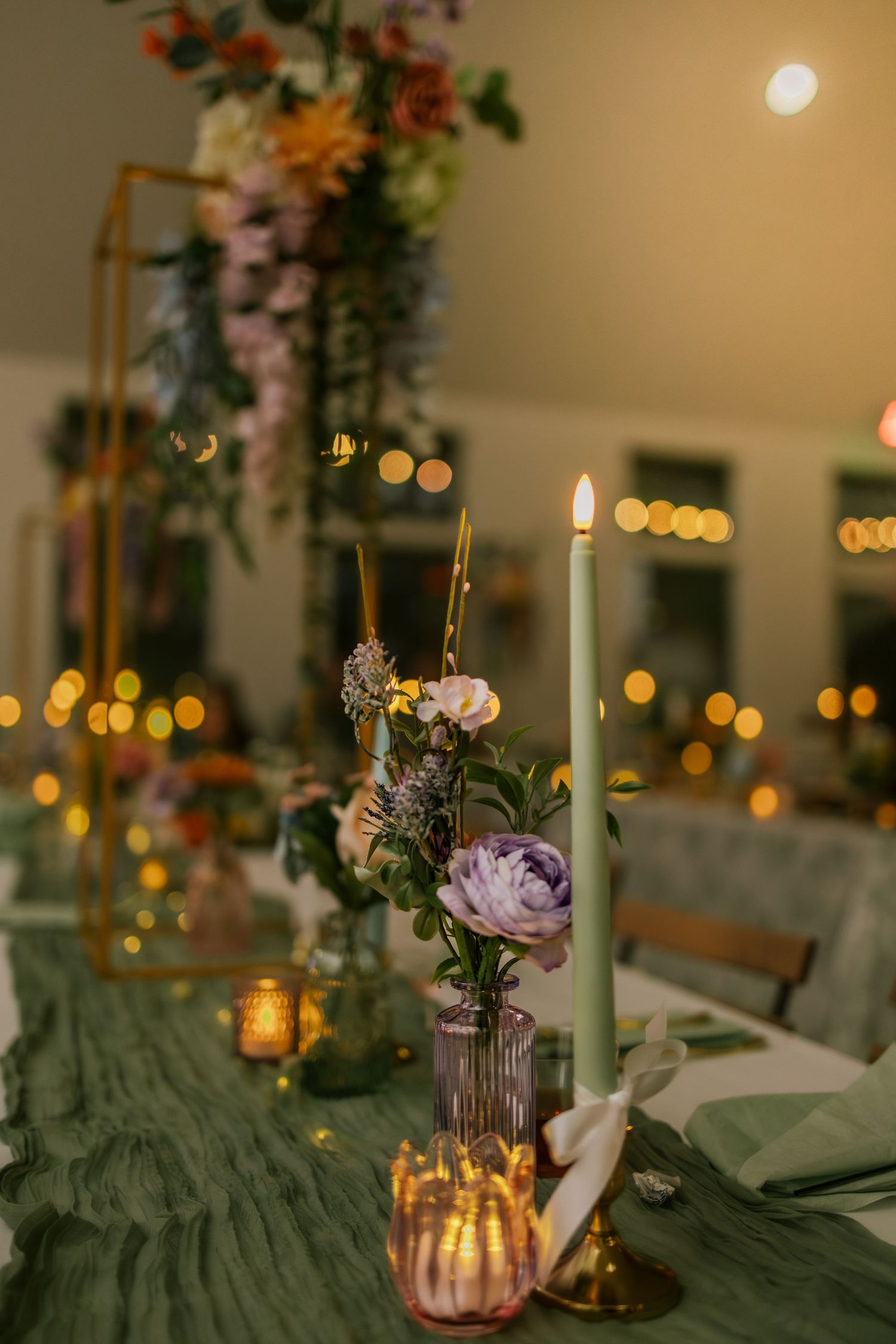 Elegant wedding table with green candles, purple floral arrangements, and warm bokeh lights in a softly lit reception venue.