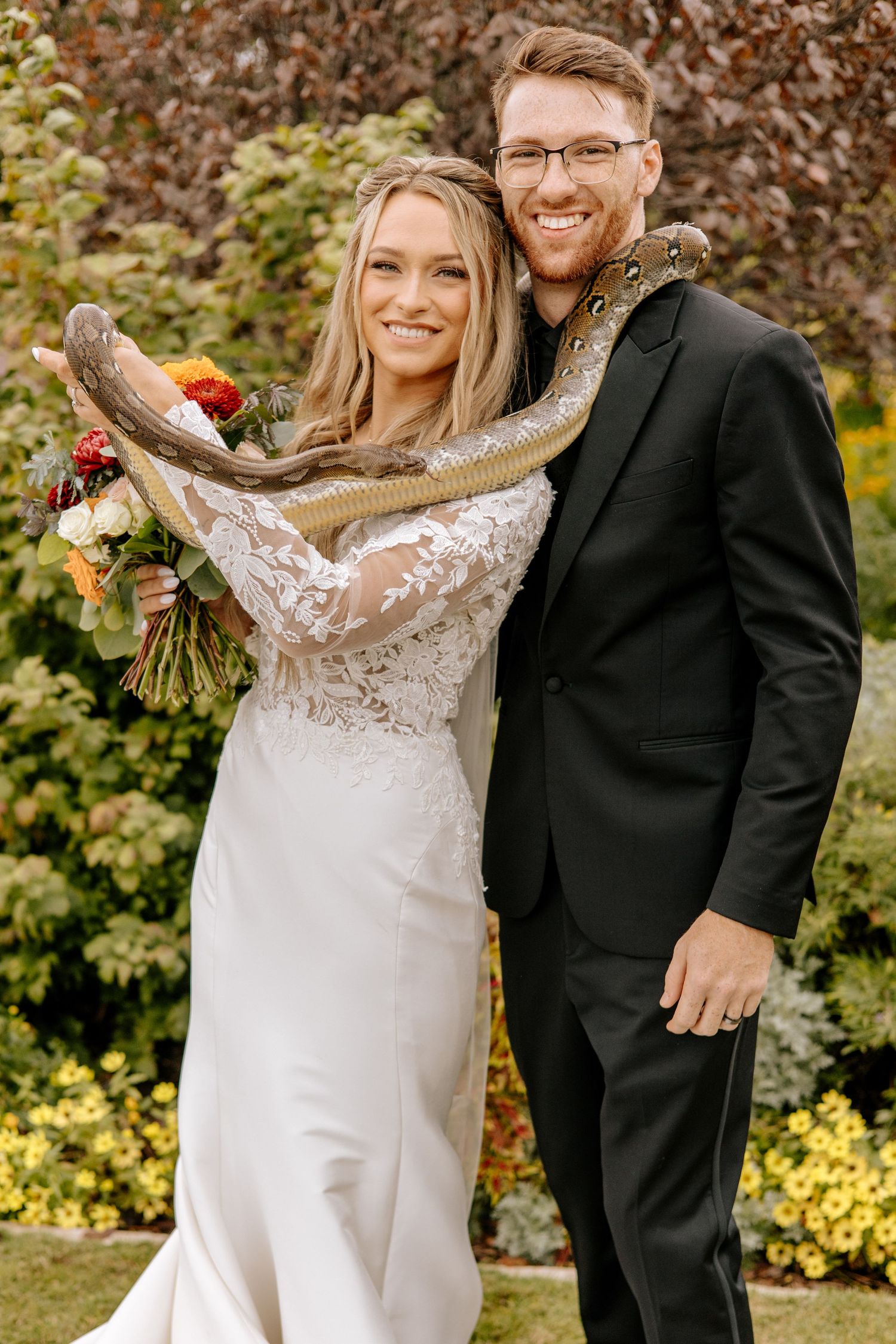 Smiling bride and groom pose with large snake draped over shoulders in outdoor garden wedding portrait.