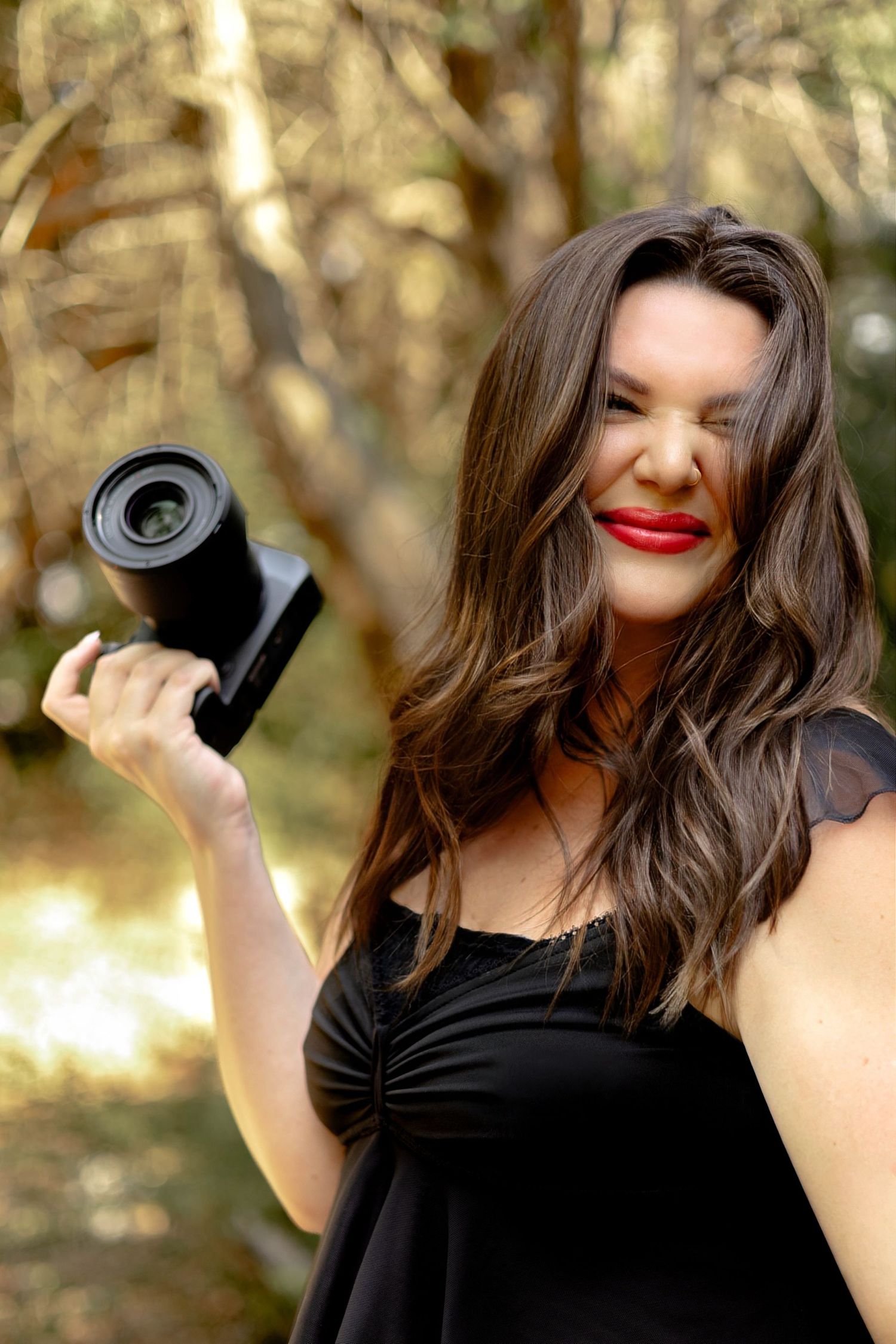A woman with long brown hair and red lips holds a black camera in a sunlit forest, wearing a black dress.