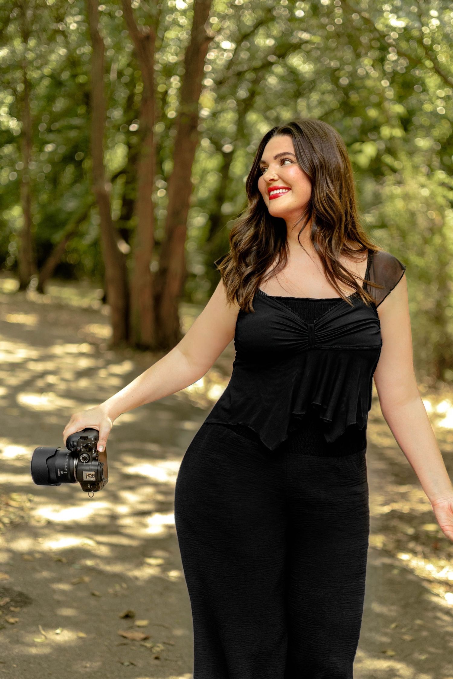 Woman in black dress holding DSLR camera smiling outdoors in lush green forest setting.