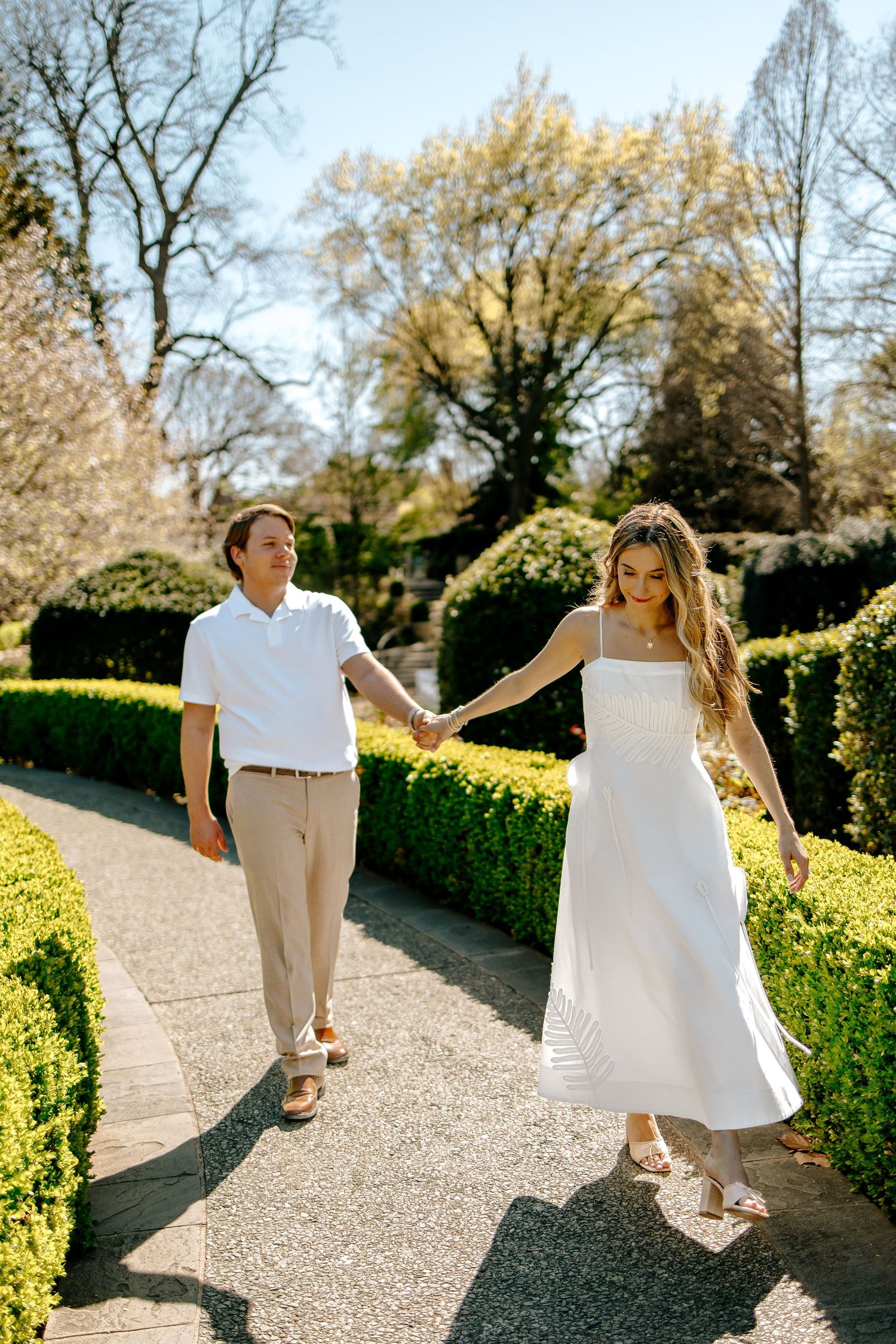 Couple holding hands walking along a garden path lined with manicured hedges on a sunny day.