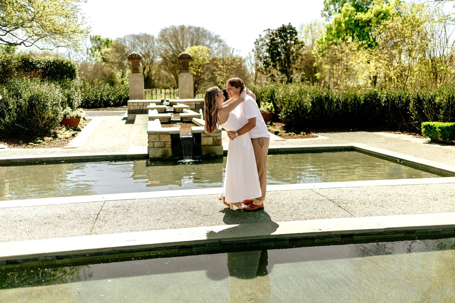 Couple sharing a romantic kiss in white outfits beside a reflective garden fountain surrounded by lush greenery.