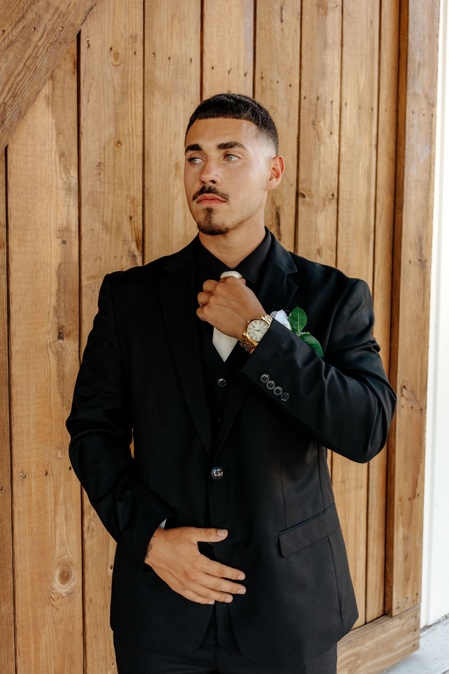 Groom in black suit adjusts tie while wearing gold watch against wooden barn wall.