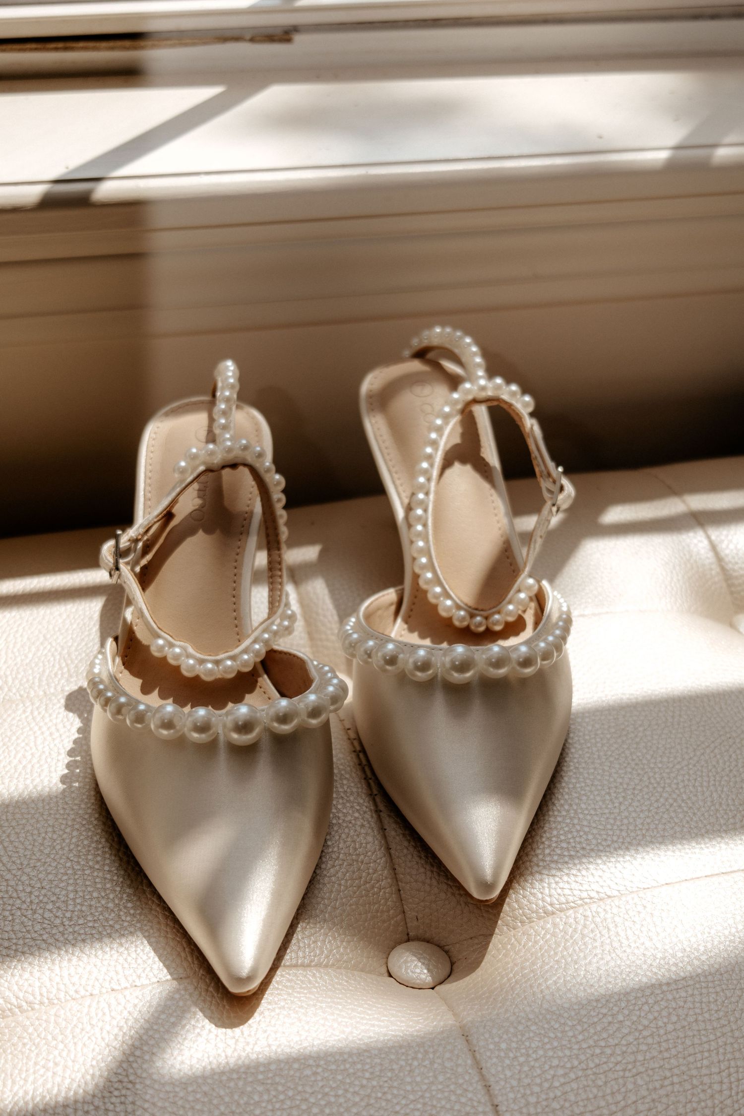 Elegant pearl-embellished satin bridal heels resting on a white leather surface in warm sunlight.