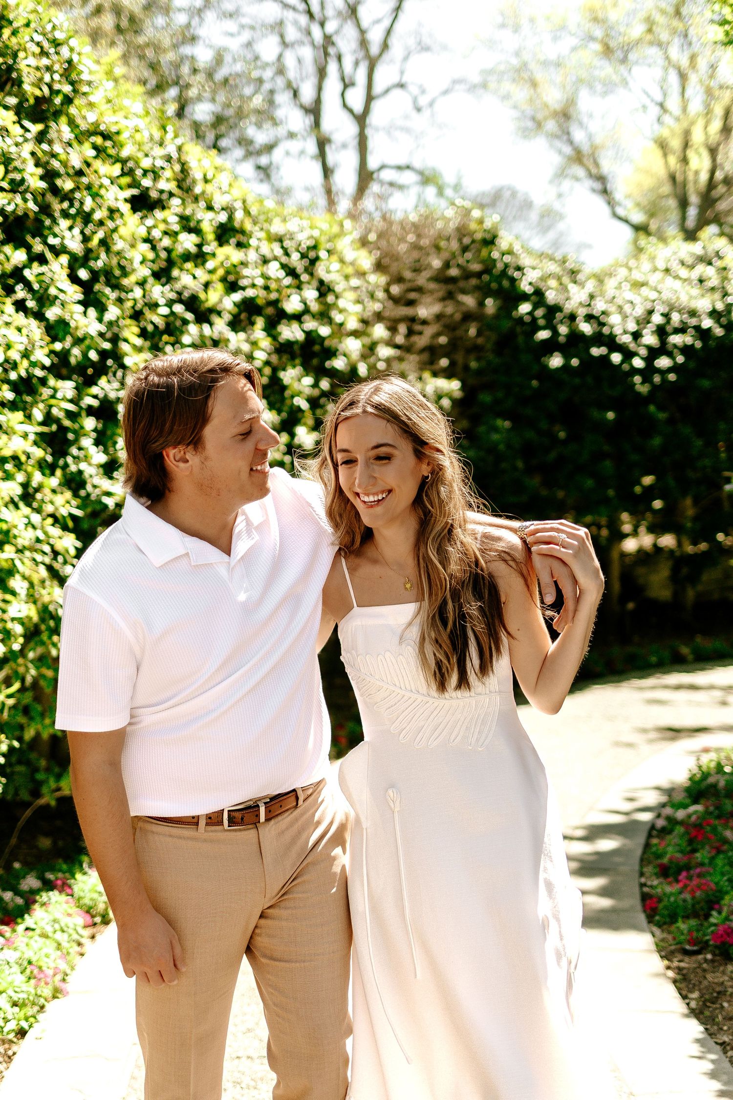 A smiling couple walks together in a lush garden, the woman in a white dress and man in a pink polo shirt.