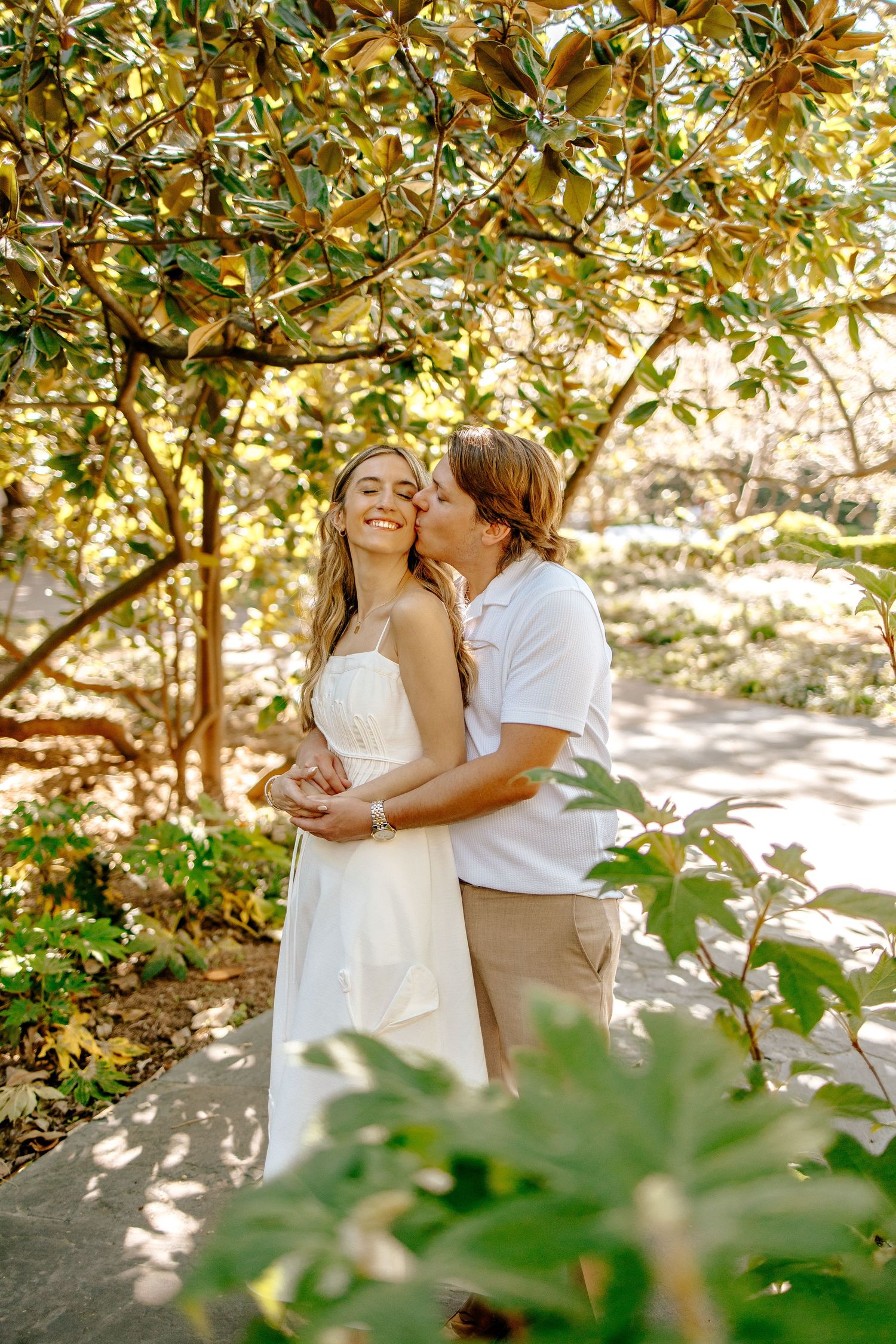 Man kissing smiling woman on the cheek while embracing under lush magnolia trees in a garden.