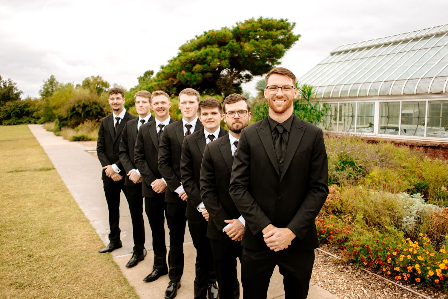 Groom leads groomsmen in matching black suits along garden path at botanical garden venue.