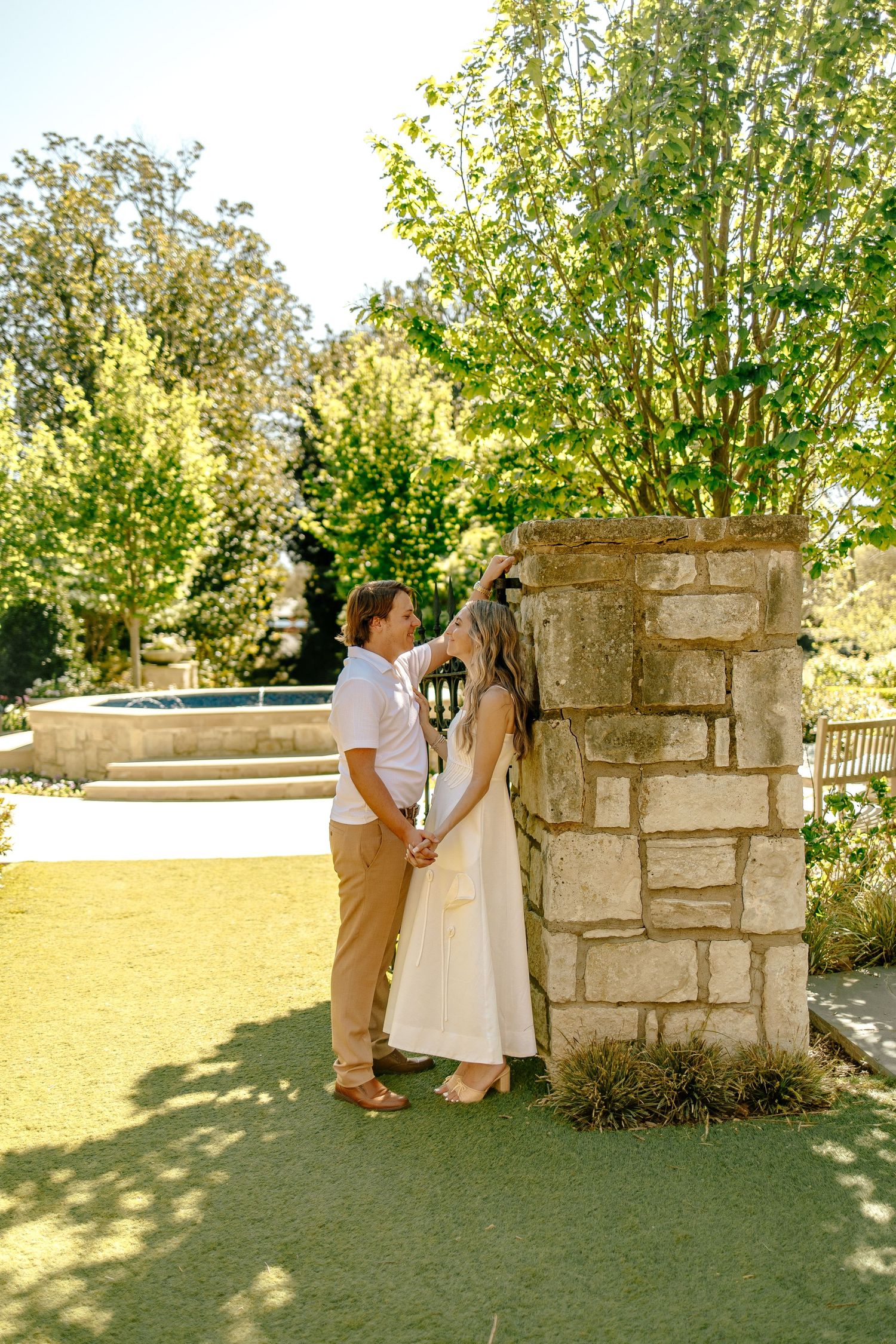 Couple holding hands beside a stone pillar with a fountain and green lawn in the background.