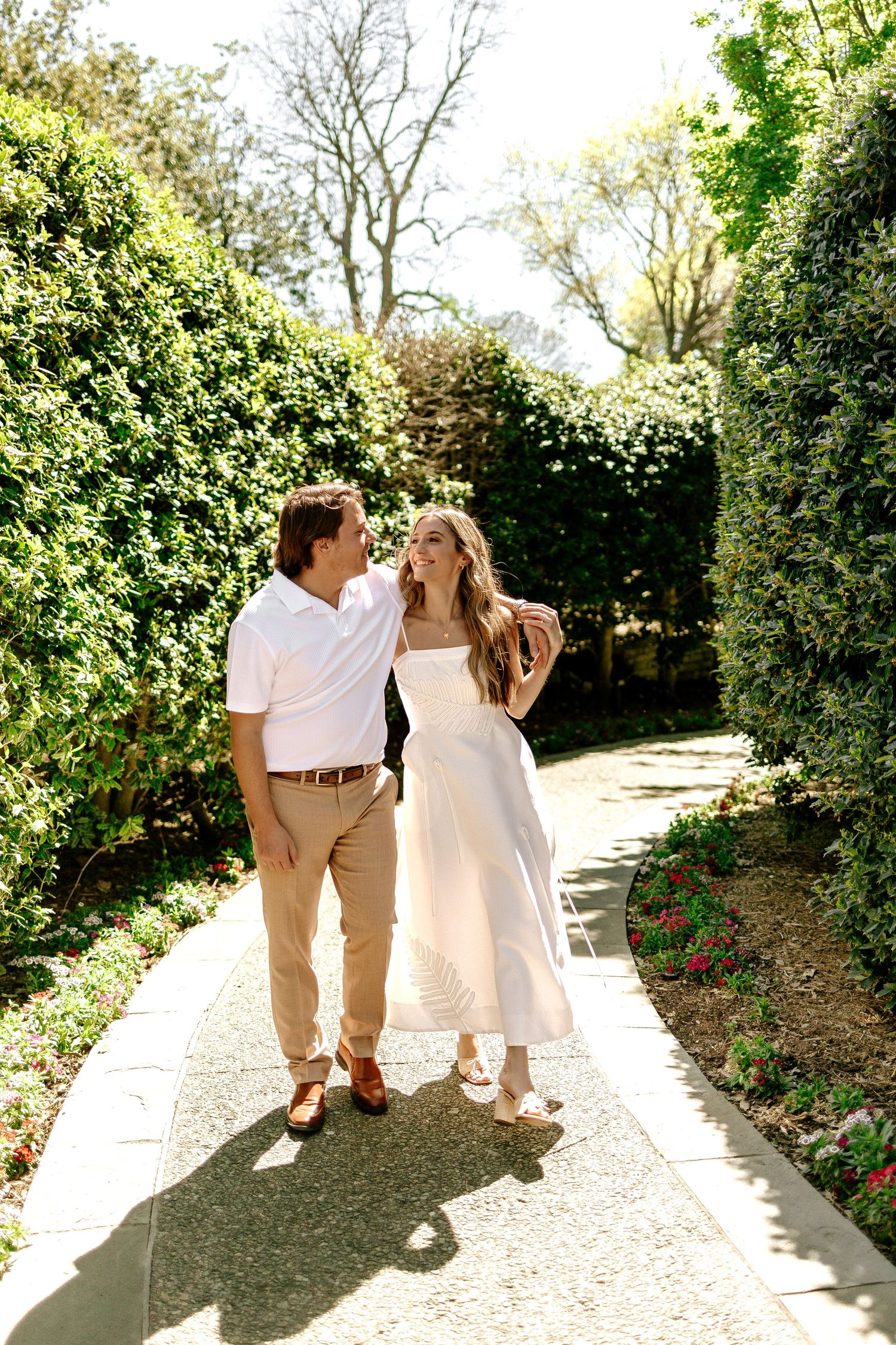 Couple walking hand in hand on a garden path surrounded by lush green hedges on a sunny day.