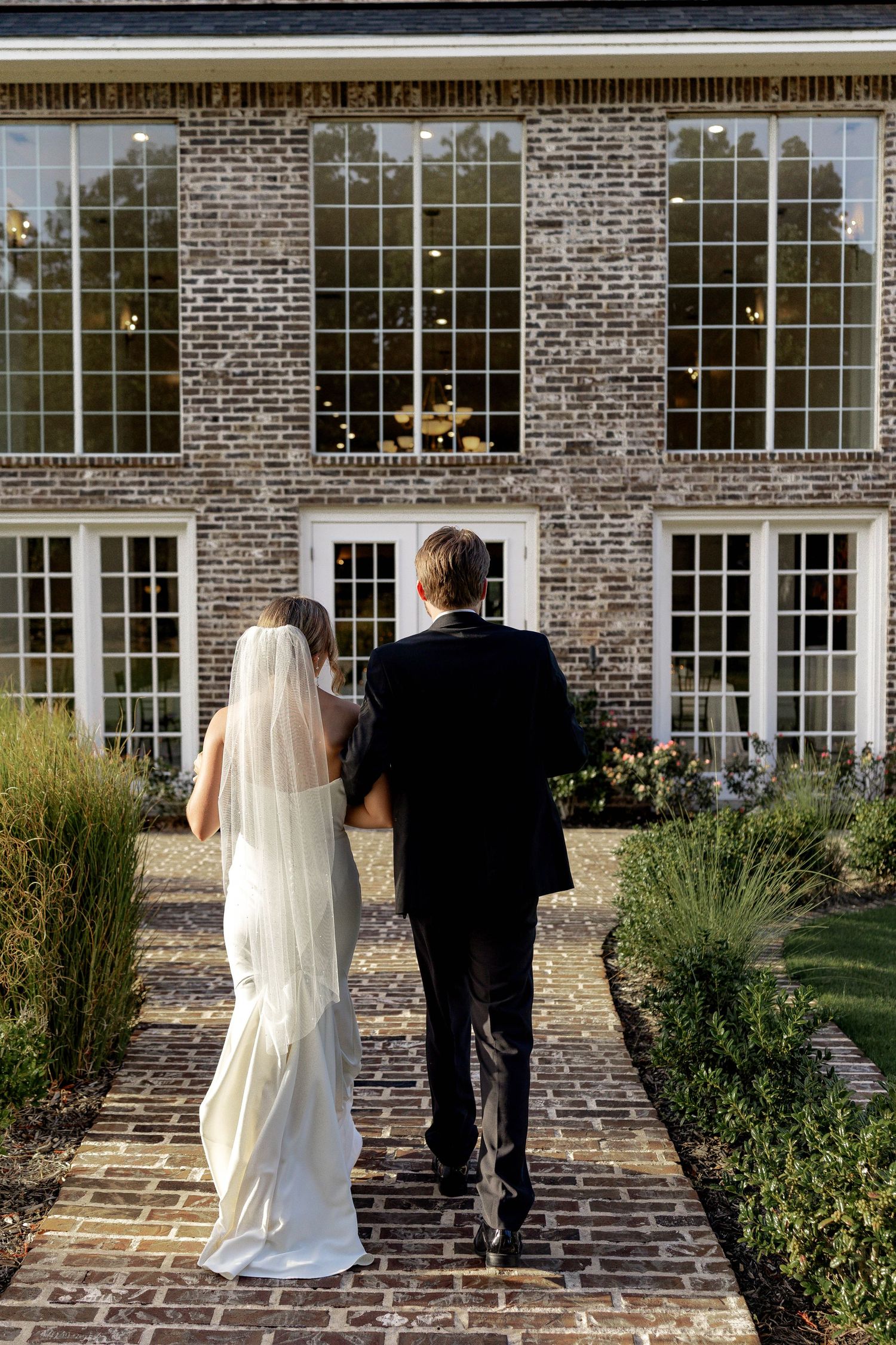 Bride and groom walking hand in hand toward elegant brick mansion on a cobblestone path.