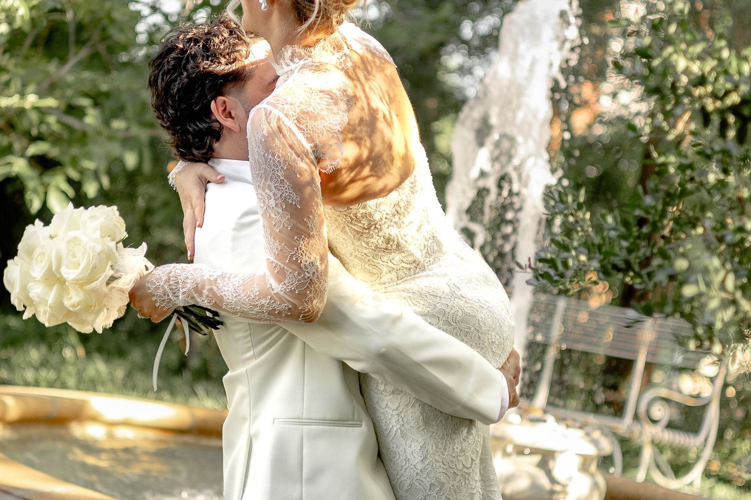 Groom lifting bride in lace wedding dress near fountain, sharing romantic kiss outdoors on wedding day.