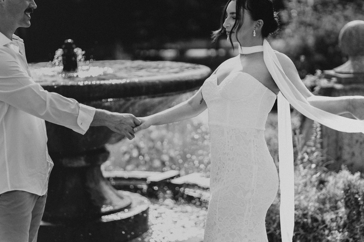 Black and white photo of a bride and groom holding hands near a fountain, bride wearing elegant gown with flowing veil.