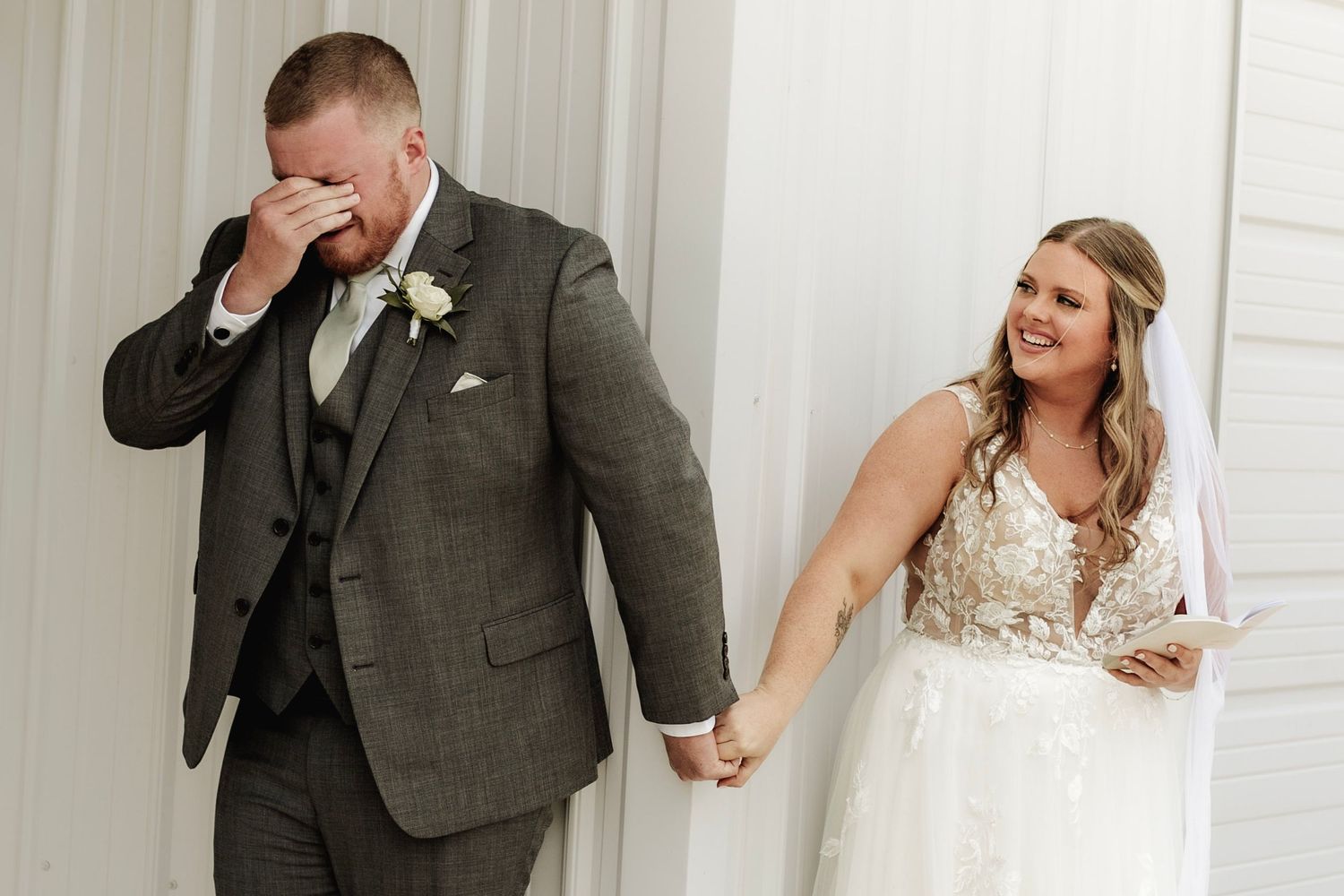 Emotional groom wiping tears while smiling bride holds his hand during wedding ceremony in white venue.