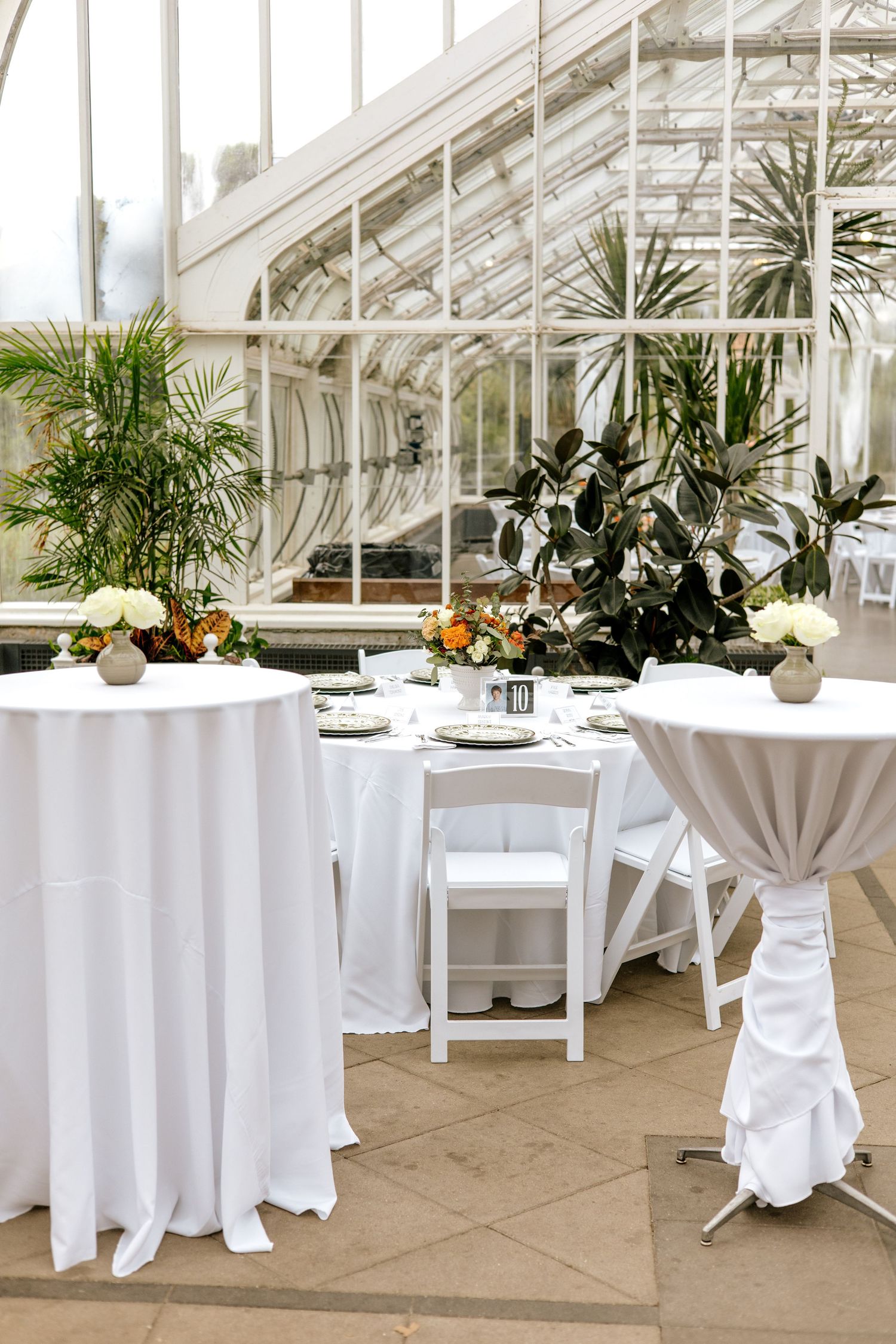 Wedding reception setup in glass greenhouse with white draped tables, tropical plants, and orange floral centerpieces.