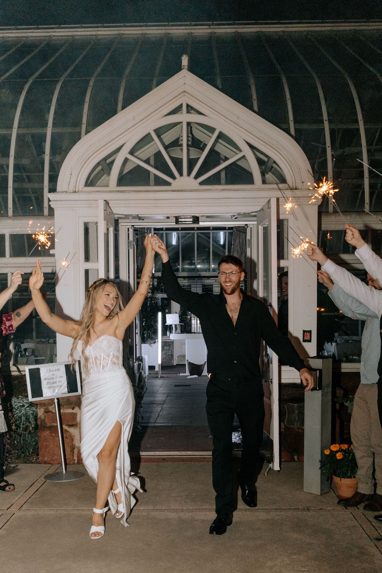 Bride and groom exit wedding venue through sparkler send-off at elegant greenhouse.