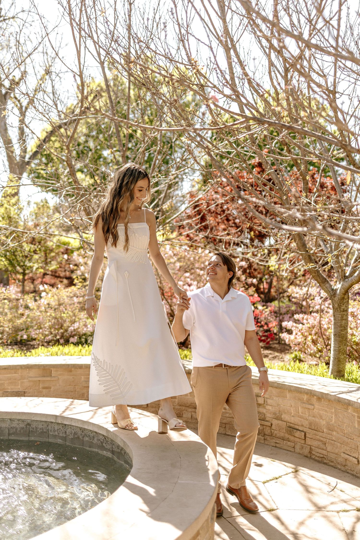 Couple holding hands near a decorative fountain surrounded by bare trees in a beautiful garden.