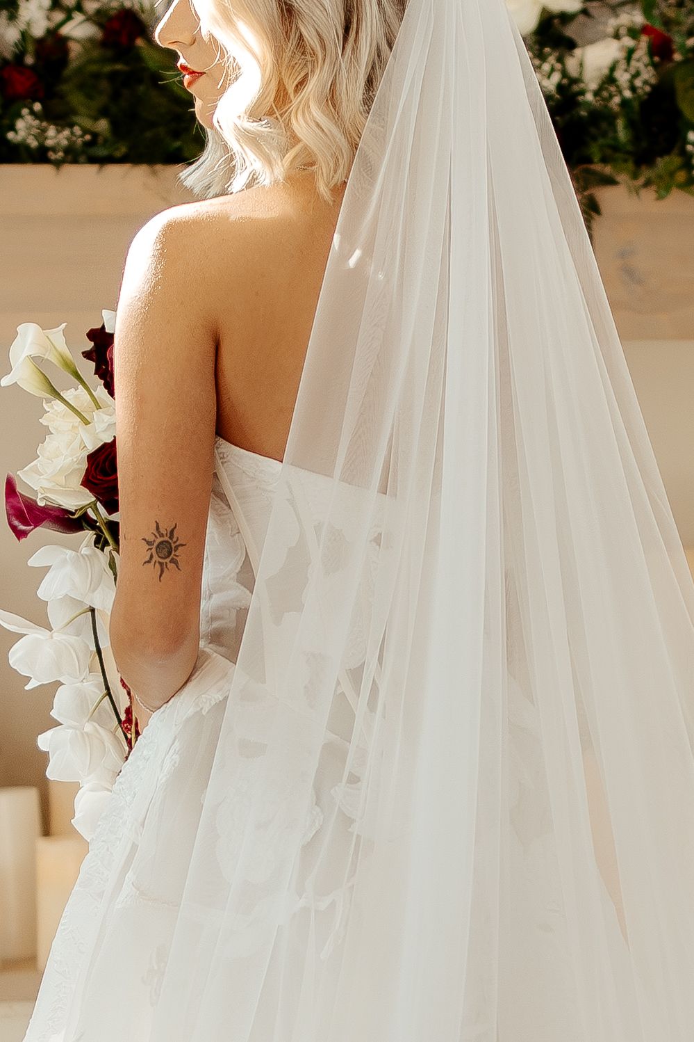 Bride in strapless white gown with veil and sun tattoo on arm holds bouquet of white and burgundy flowers.