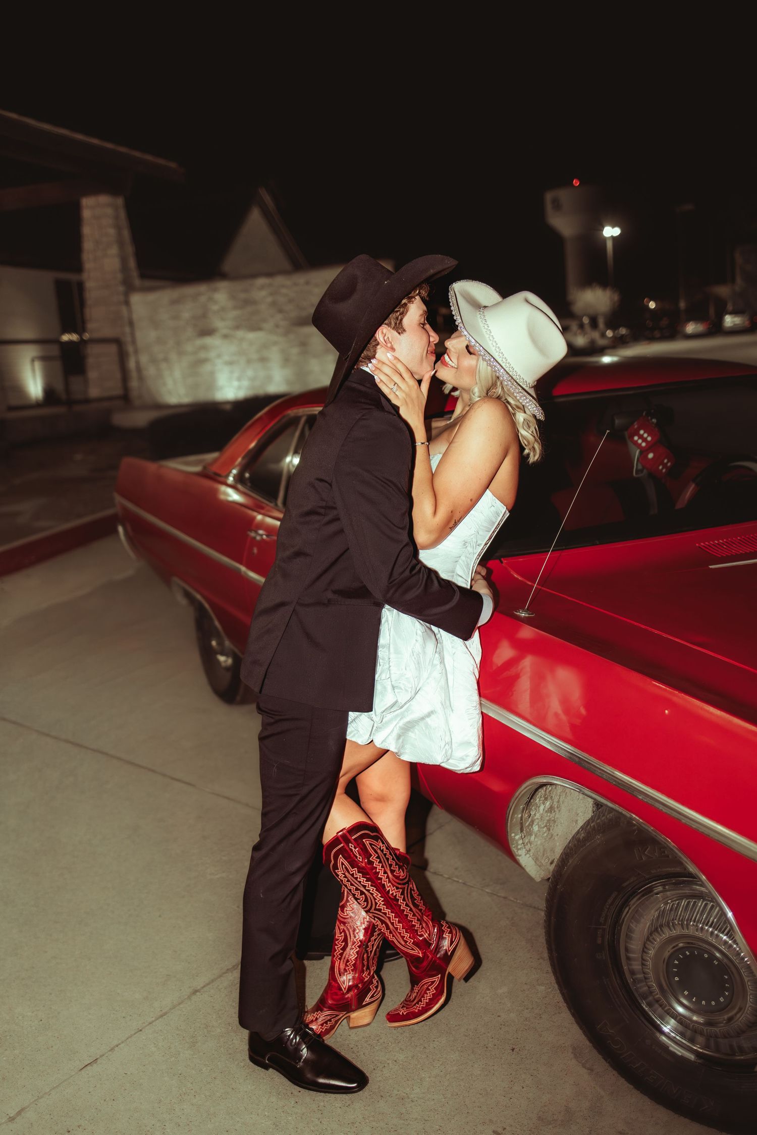 Couple in cowboy hats kissing beside a red classic car at night, wearing western boots and formal attire.