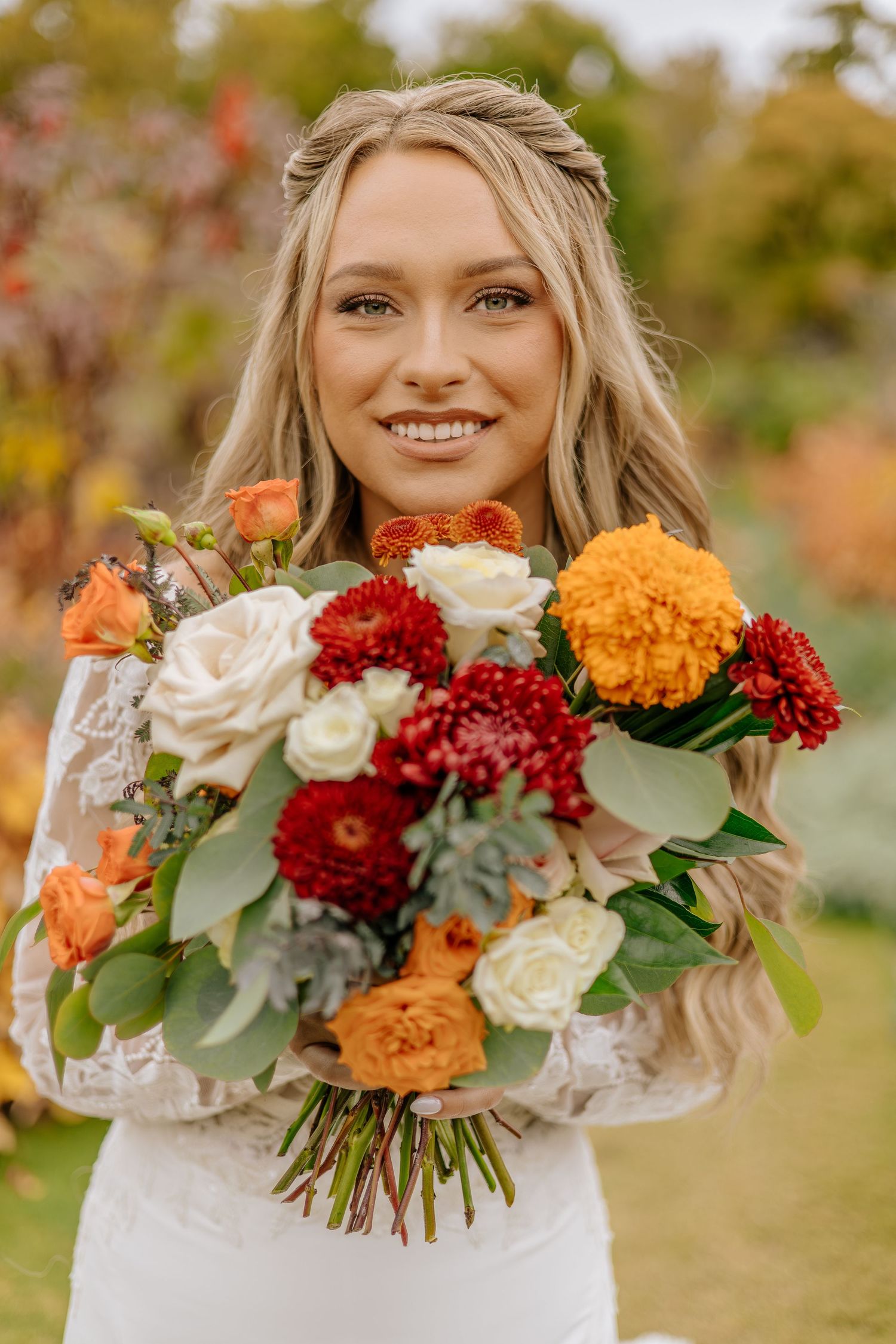 Radiant bride holds a lush autumn bouquet featuring orange marigolds, red dahlias, and white roses in a garden setting.