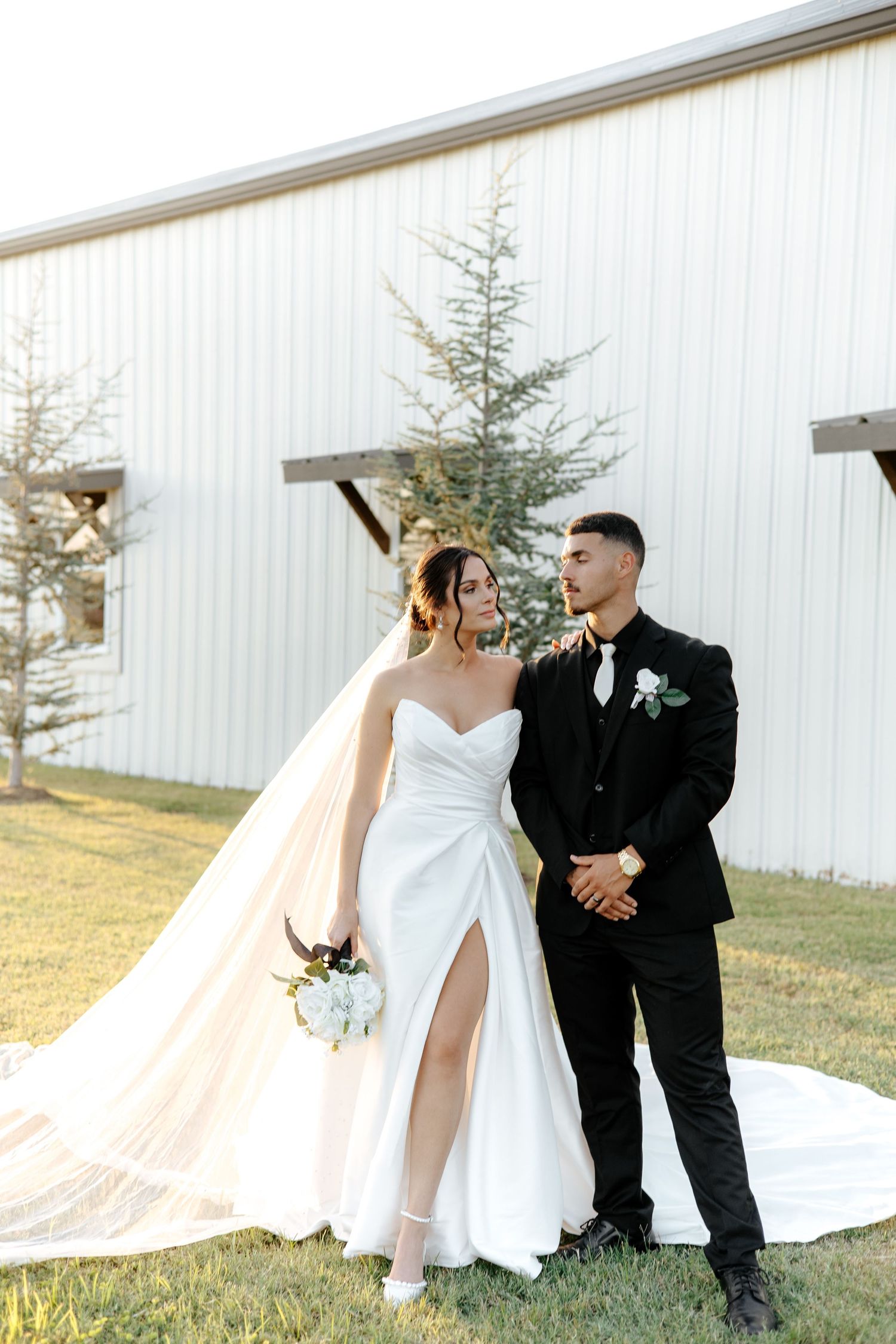 Bride in strapless gown with flowing veil and groom in black suit pose at barn venue.