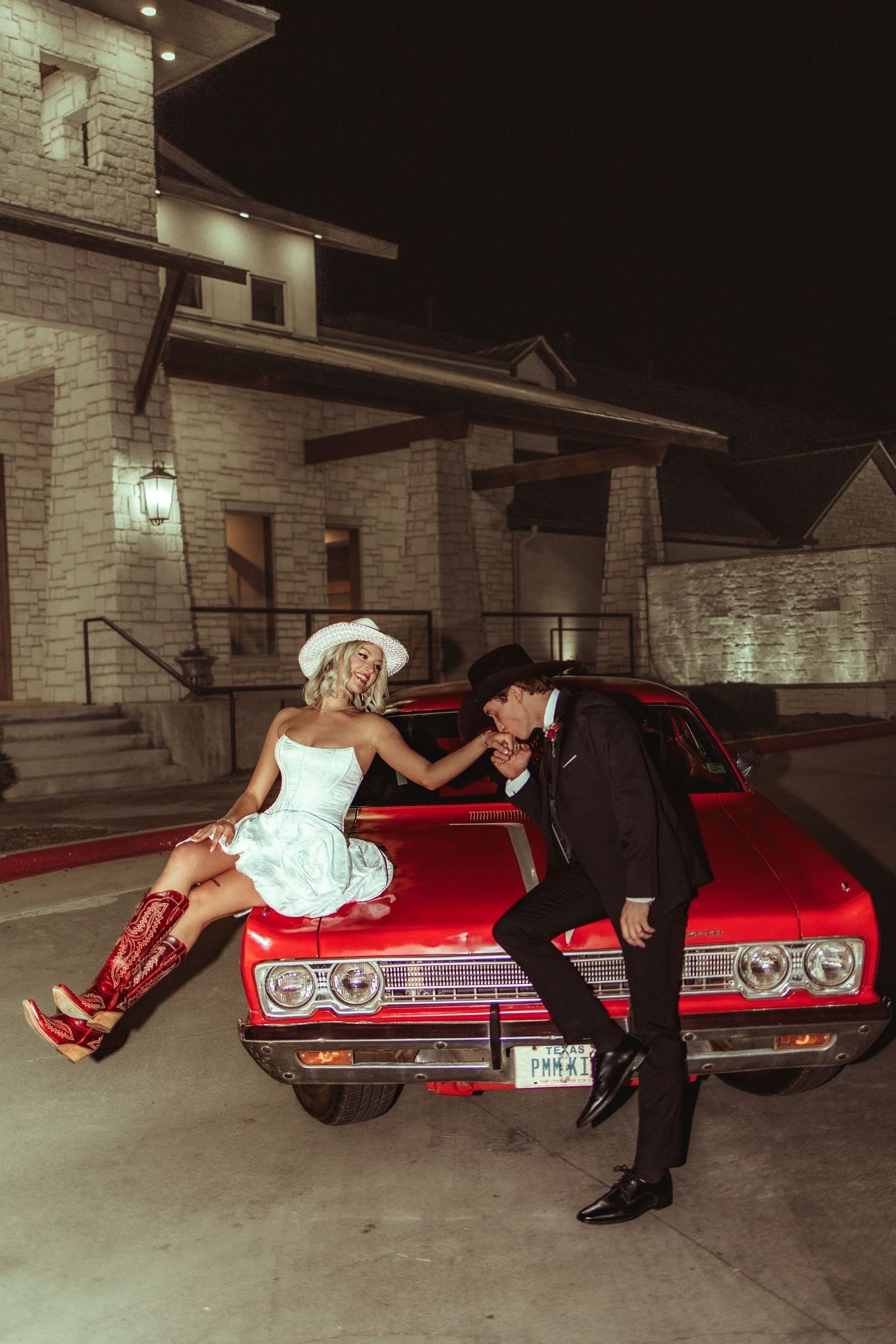 Couple in western attire poses by a red classic car at night, bride in white dress and red boots, groom in black tuxedo.