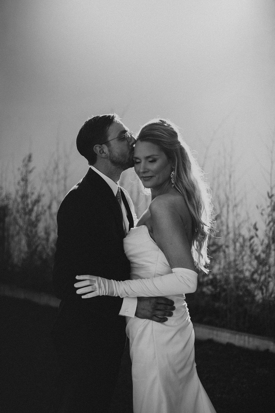 Black and white wedding photo of groom kissing bride's cheek while she smiles, wearing elegant gown and long gloves.