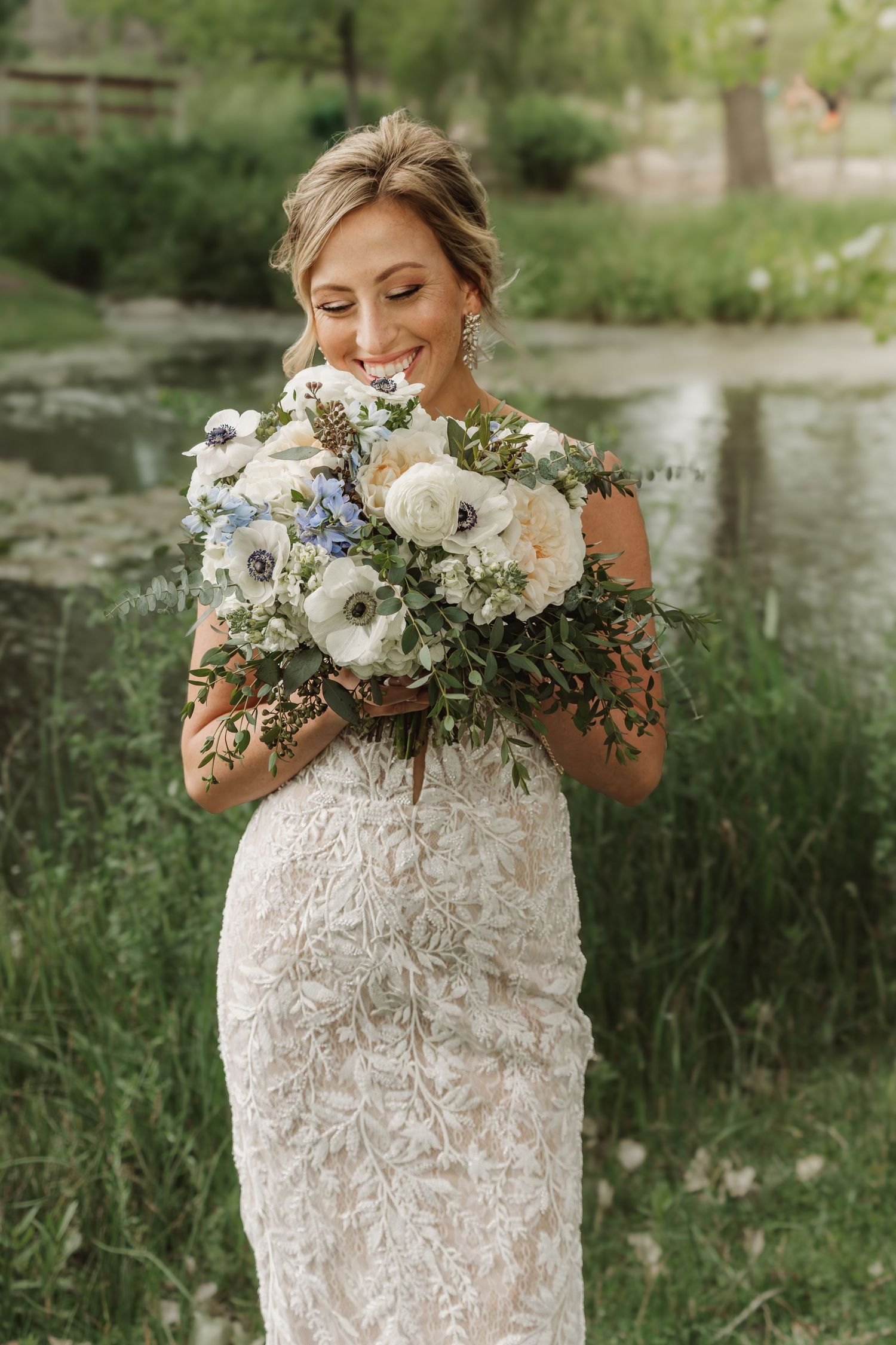 Happy bride in a fitted lace dress holding a lush white and blue bouquet near a garden pond.