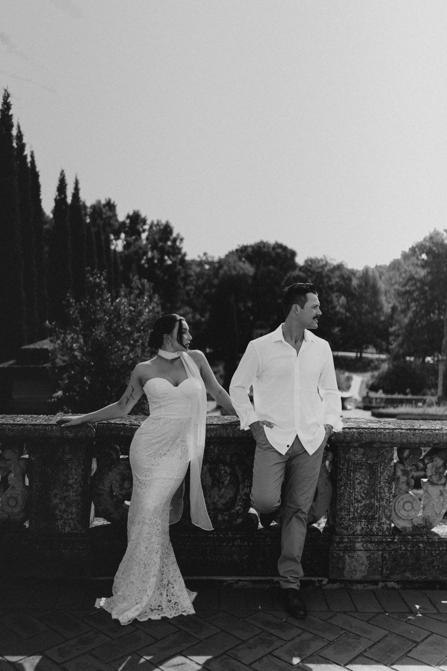 Black and white photo of a bride and groom holding hands by a stone balustrade in a lush garden setting.