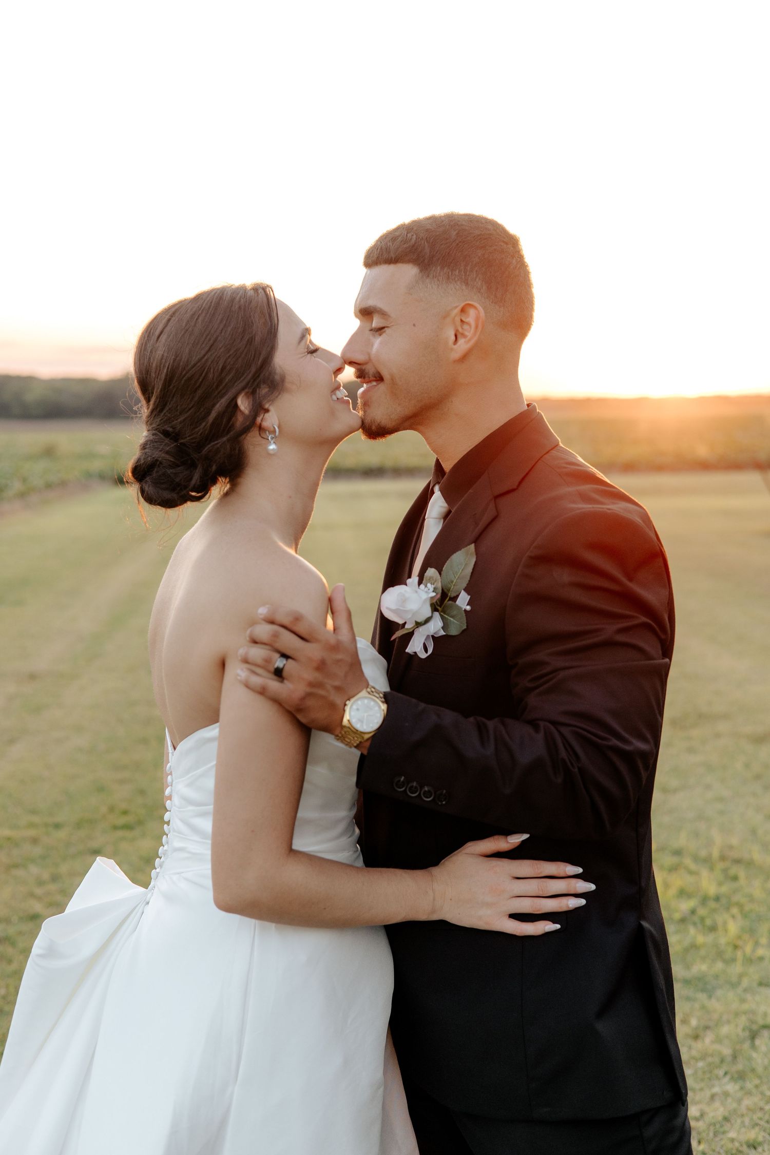 Bride and groom share a sunset kiss in an open field, groom wearing white boutonniere.