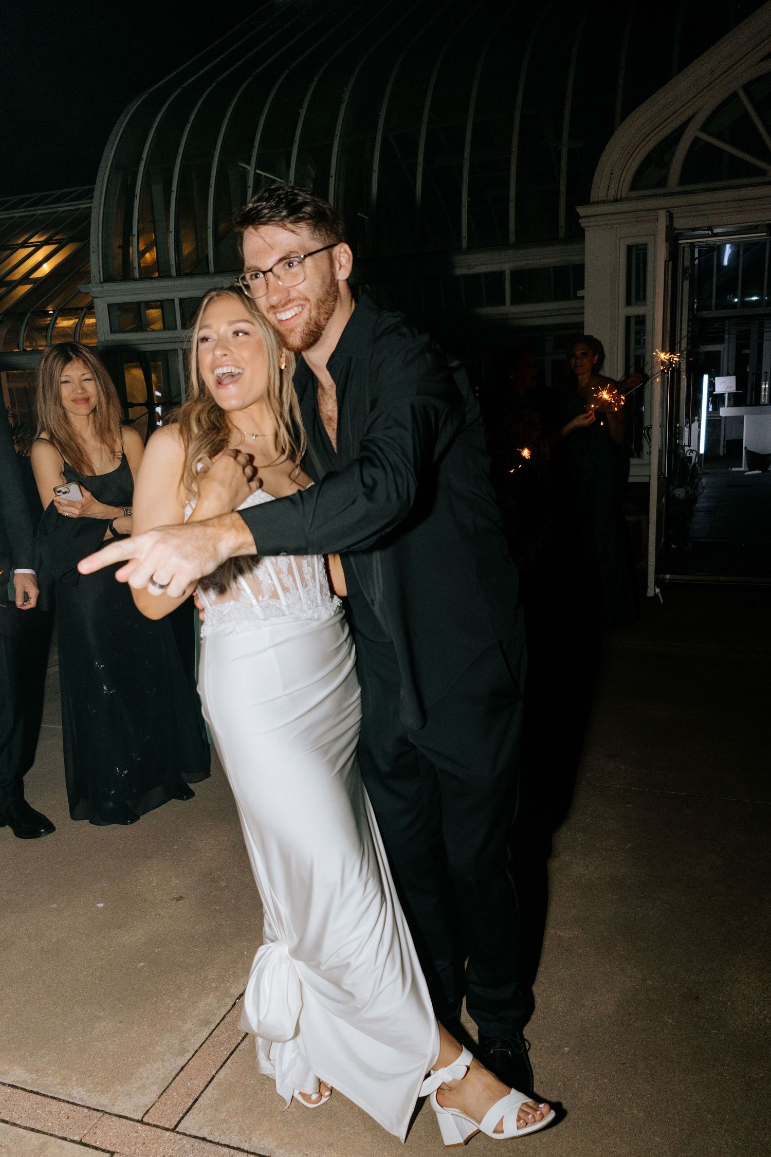 Groom in black suit embraces bride in white gown outside greenhouse wedding venue.