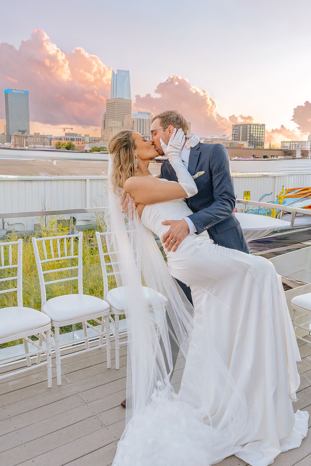 Groom dips bride for romantic kiss on rooftop wedding venue with stunning pink and purple sunset sky.