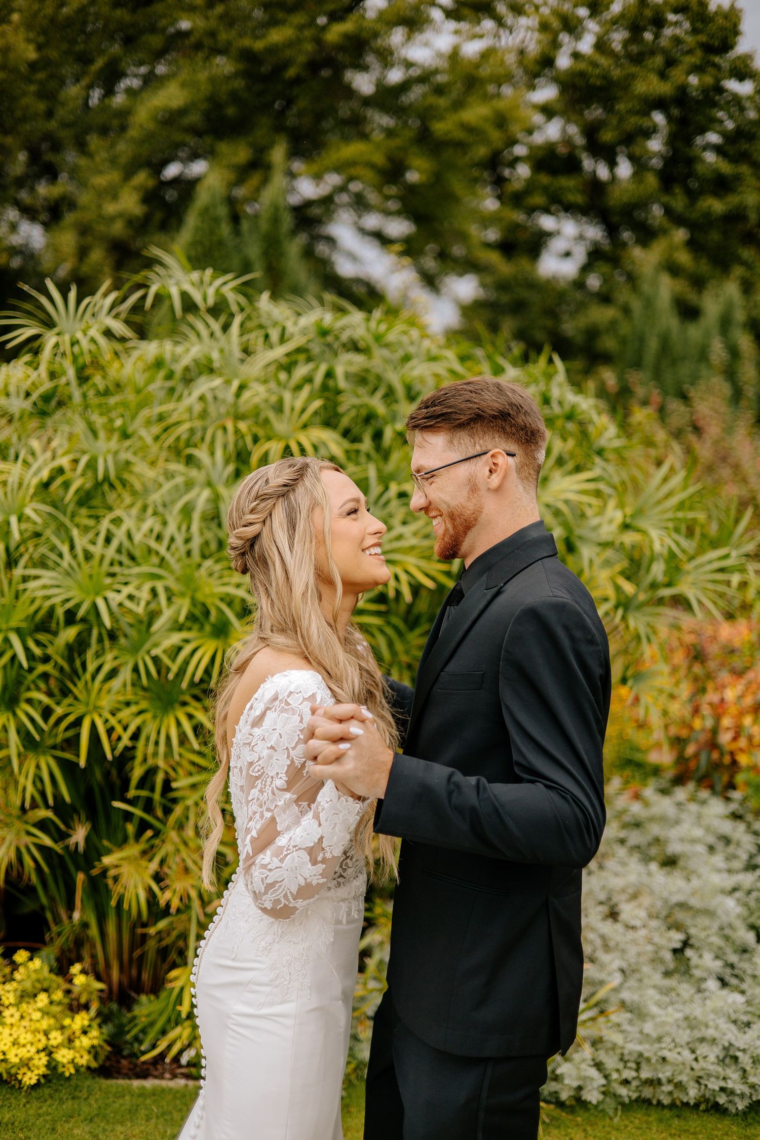 Bride and groom share a joyful moment dancing together surrounded by lush tropical garden greenery on their wedding day.