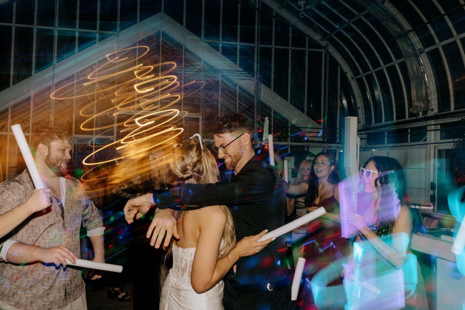 Bride and groom dancing together with guests holding glow sticks at a wedding reception.