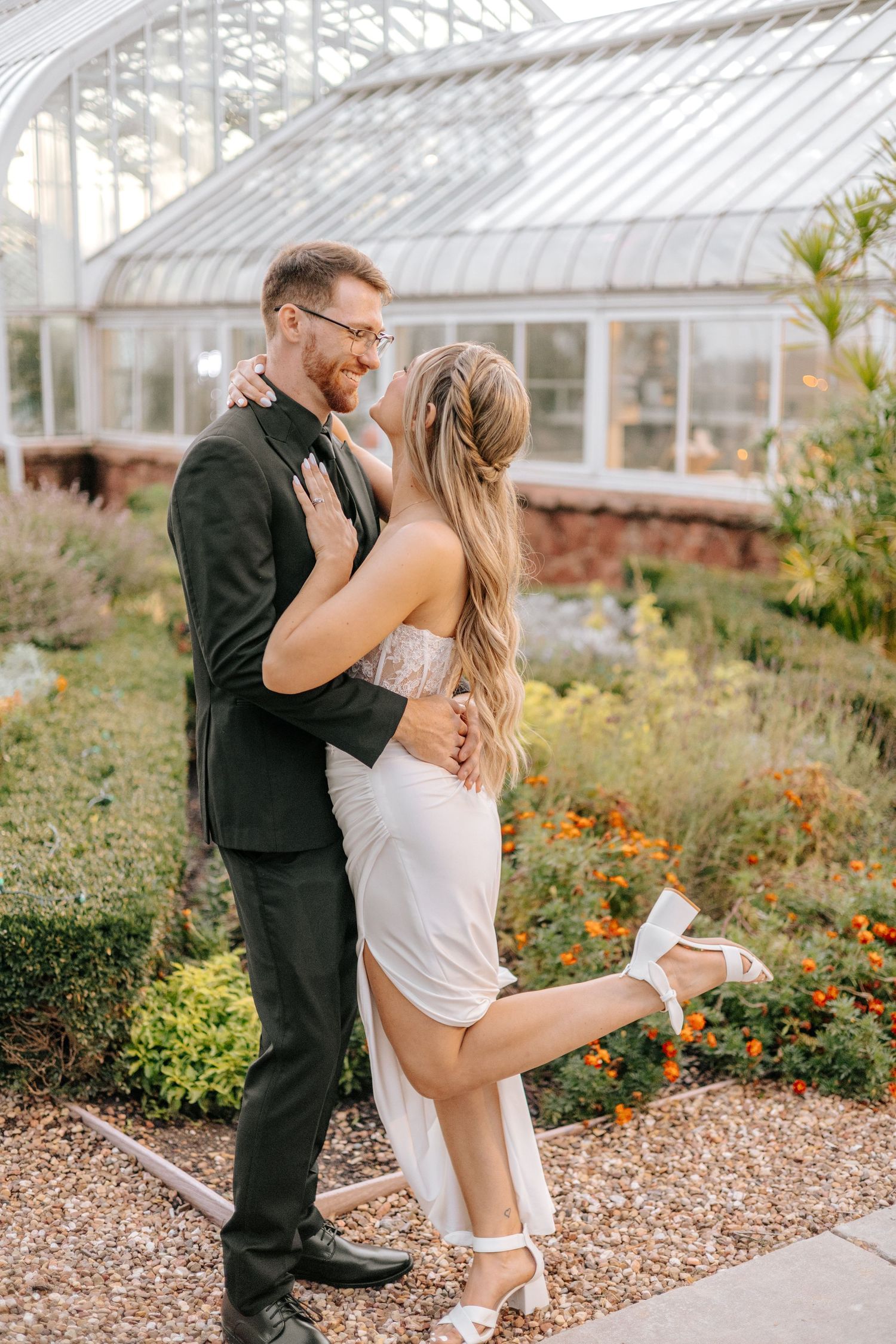 Bride kicks heel up joyfully while embracing laughing groom near glass greenhouse garden backdrop.