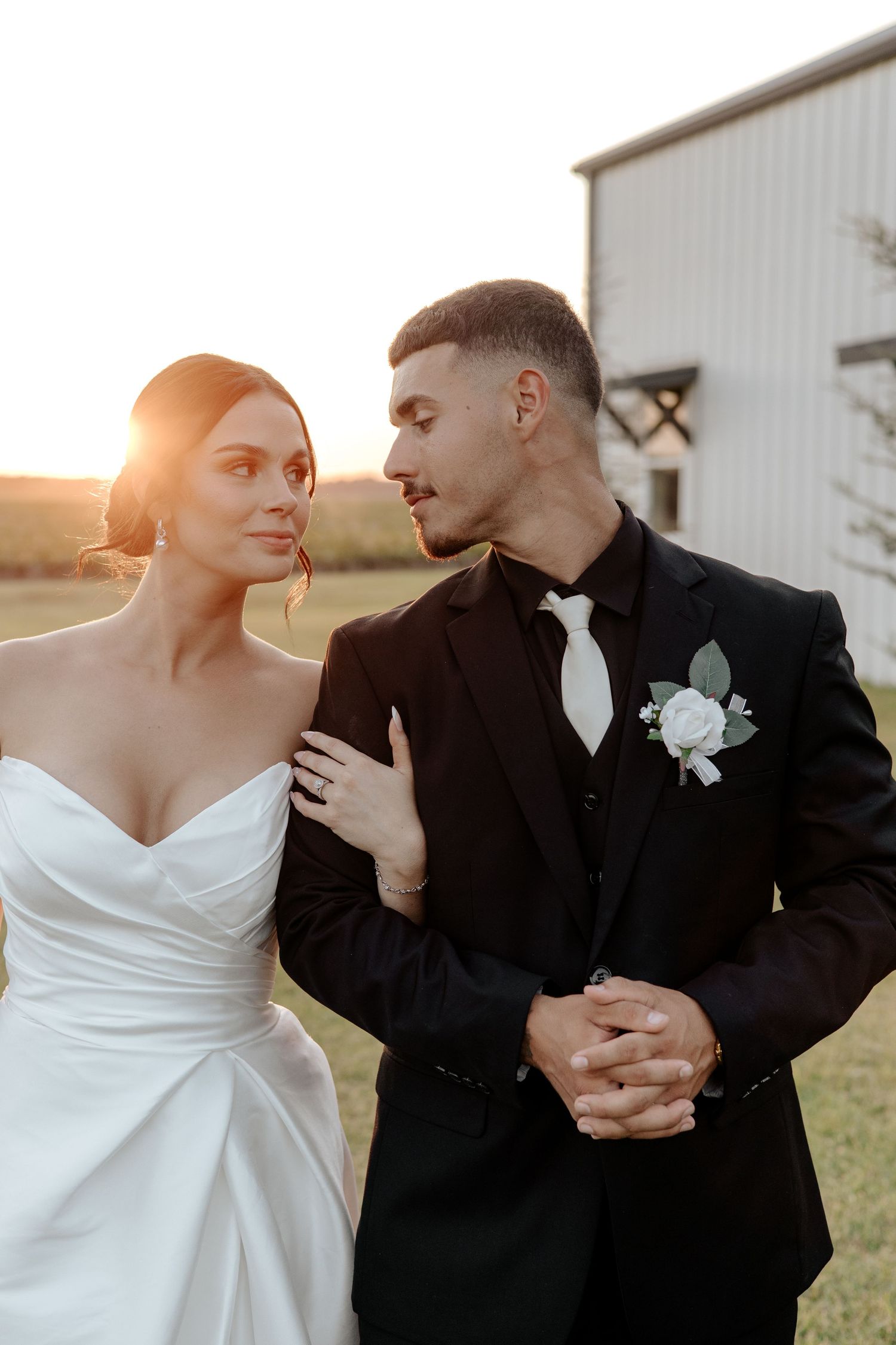 Elegant bride and groom in black tuxedo share a romantic glance at golden hour near white barn.