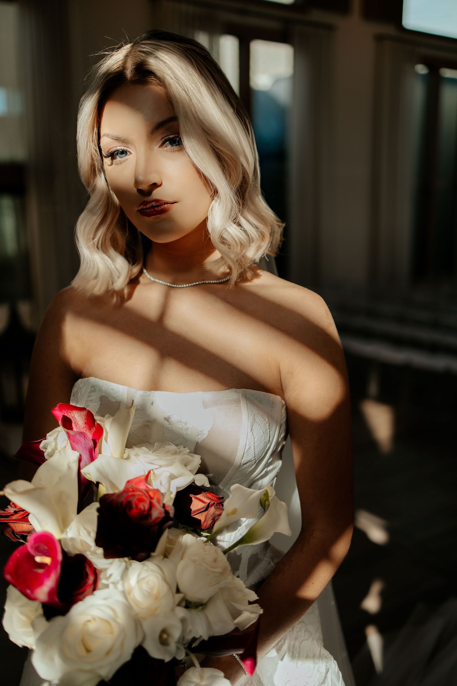 Blonde bride in strapless white gown holds colorful bouquet of roses and lilies, bathed in dramatic sunlight.