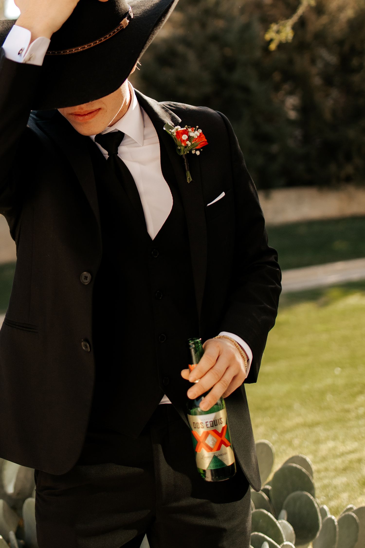 Groom in black suit and hat holding a Dos Equis beer bottle, with a red floral boutonniere, outdoors near cacti.