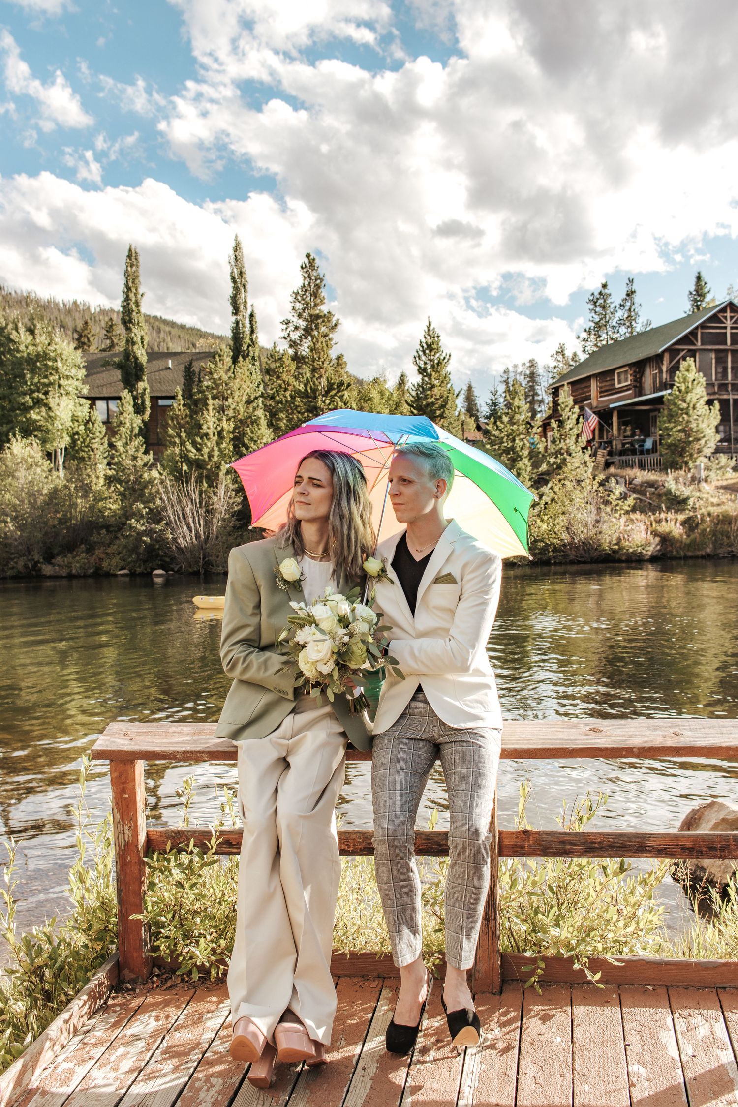 Two brides in suits share a romantic moment under a rainbow umbrella on a lakeside dock surrounded by mountain scenery.